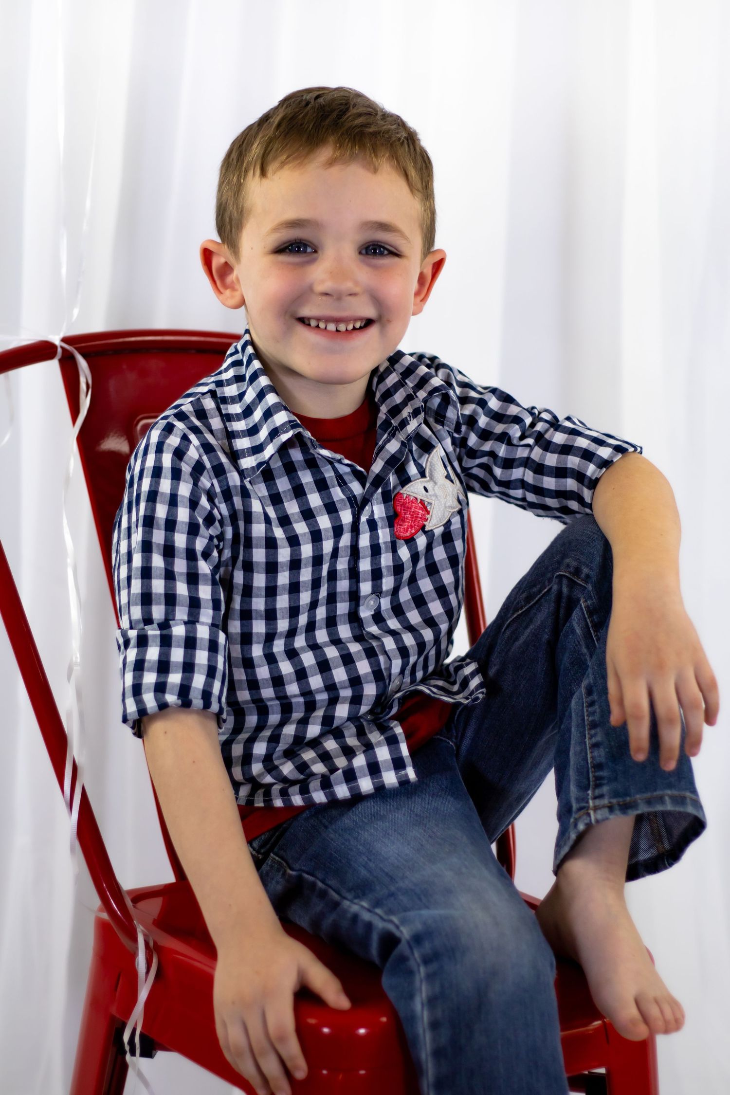 Happy young boy in blue gingham shirt and jeans sits barefoot on a red metal chair smiling.