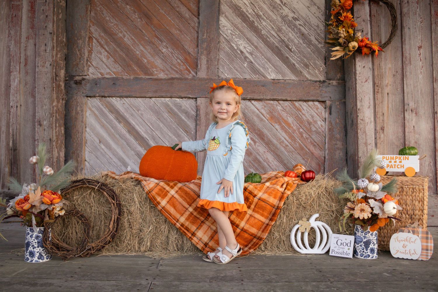 Cute little girl in blue ruffled dress sits on hay bales with pumpkins at a fall barn photo shoot.