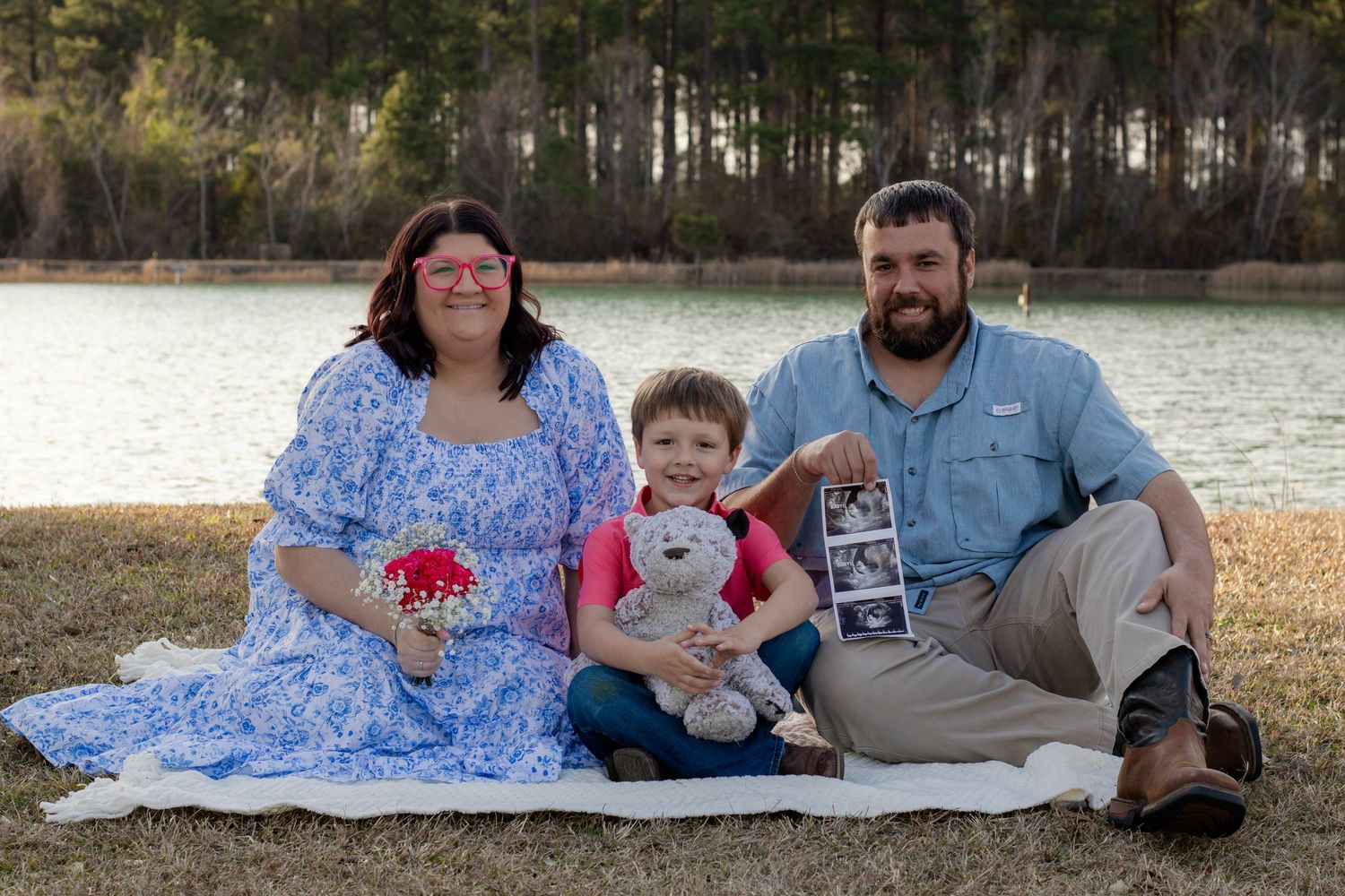 A family sits together by a lake holding flowers, a teddy bear, and ultrasound photos for a pregnancy reveal.