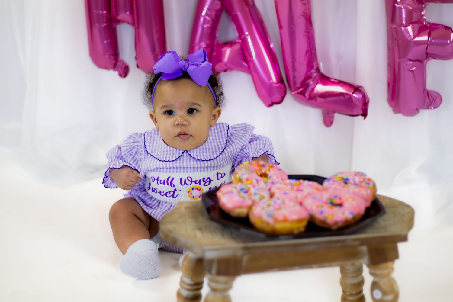 Baby girl in purple gingham romper sits beside pink sprinkle donuts at half birthday celebration.