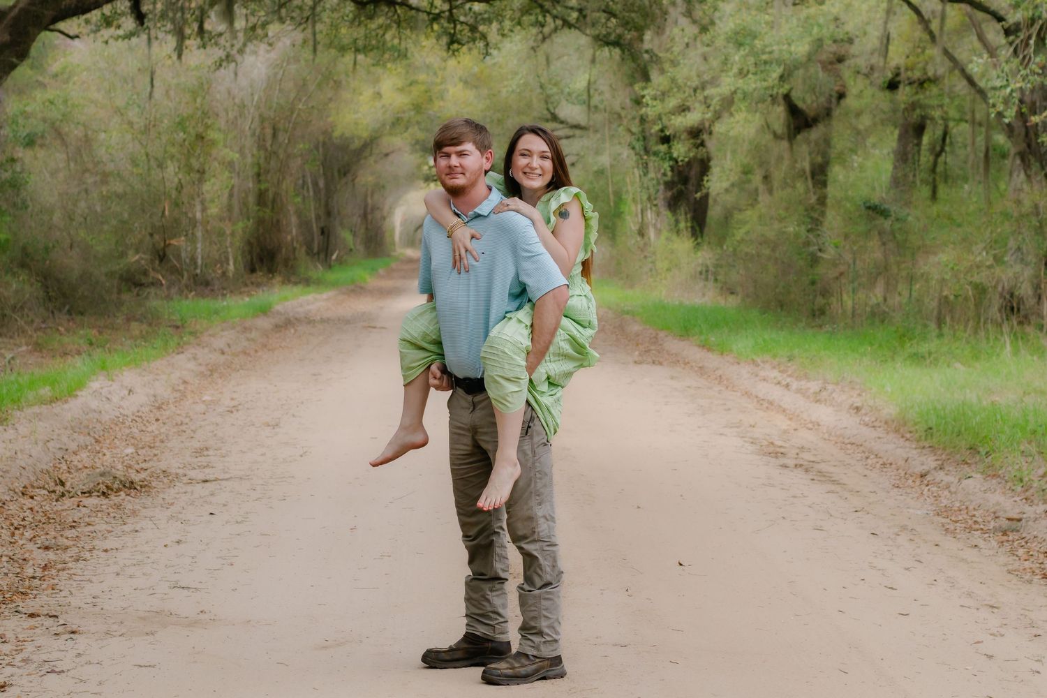 Man giving smiling woman a piggyback ride on a picturesque tree-canopied dirt road.