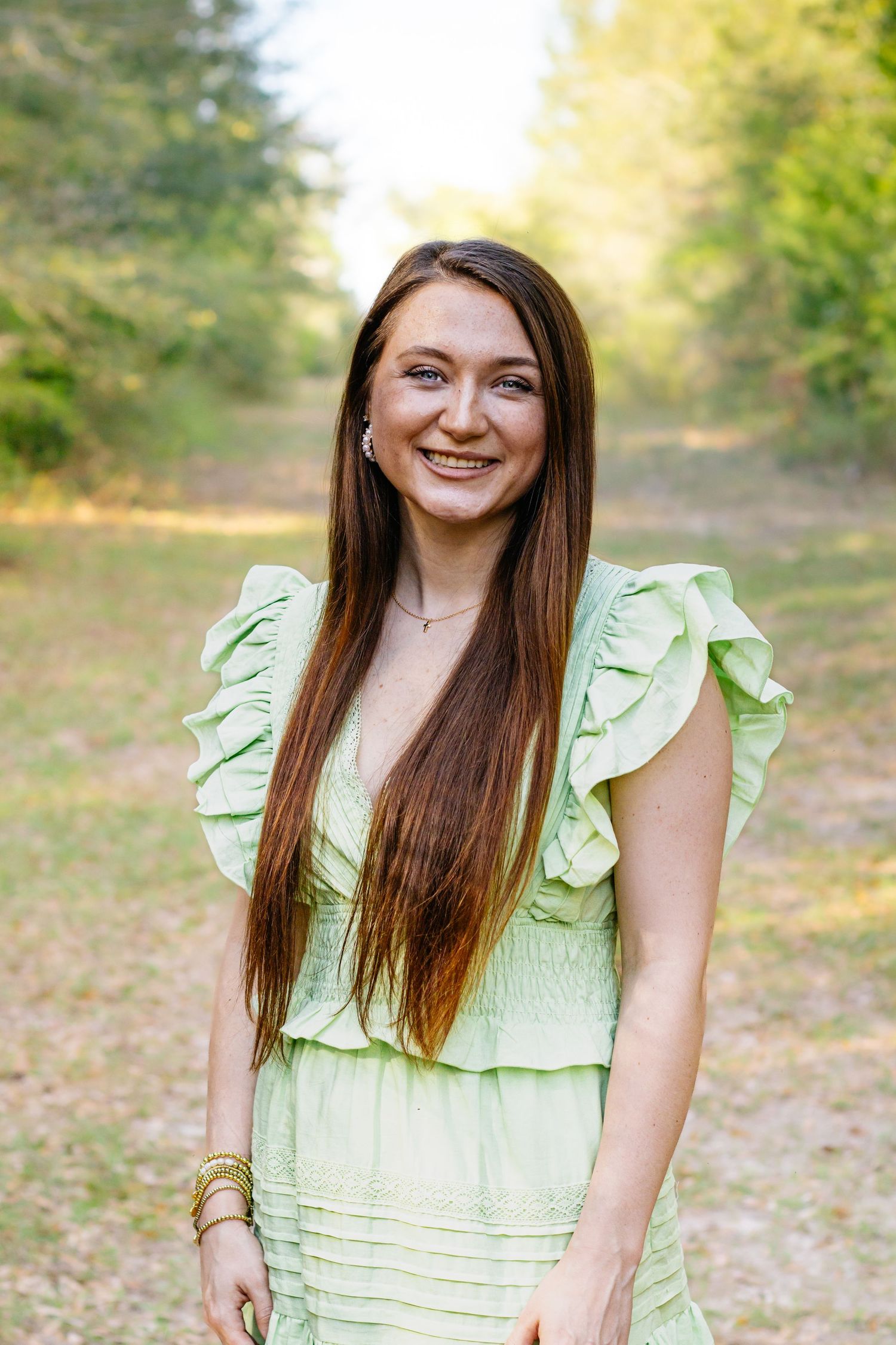 Young woman with long brown hair smiling outdoors in a green ruffled dress in a forest setting.