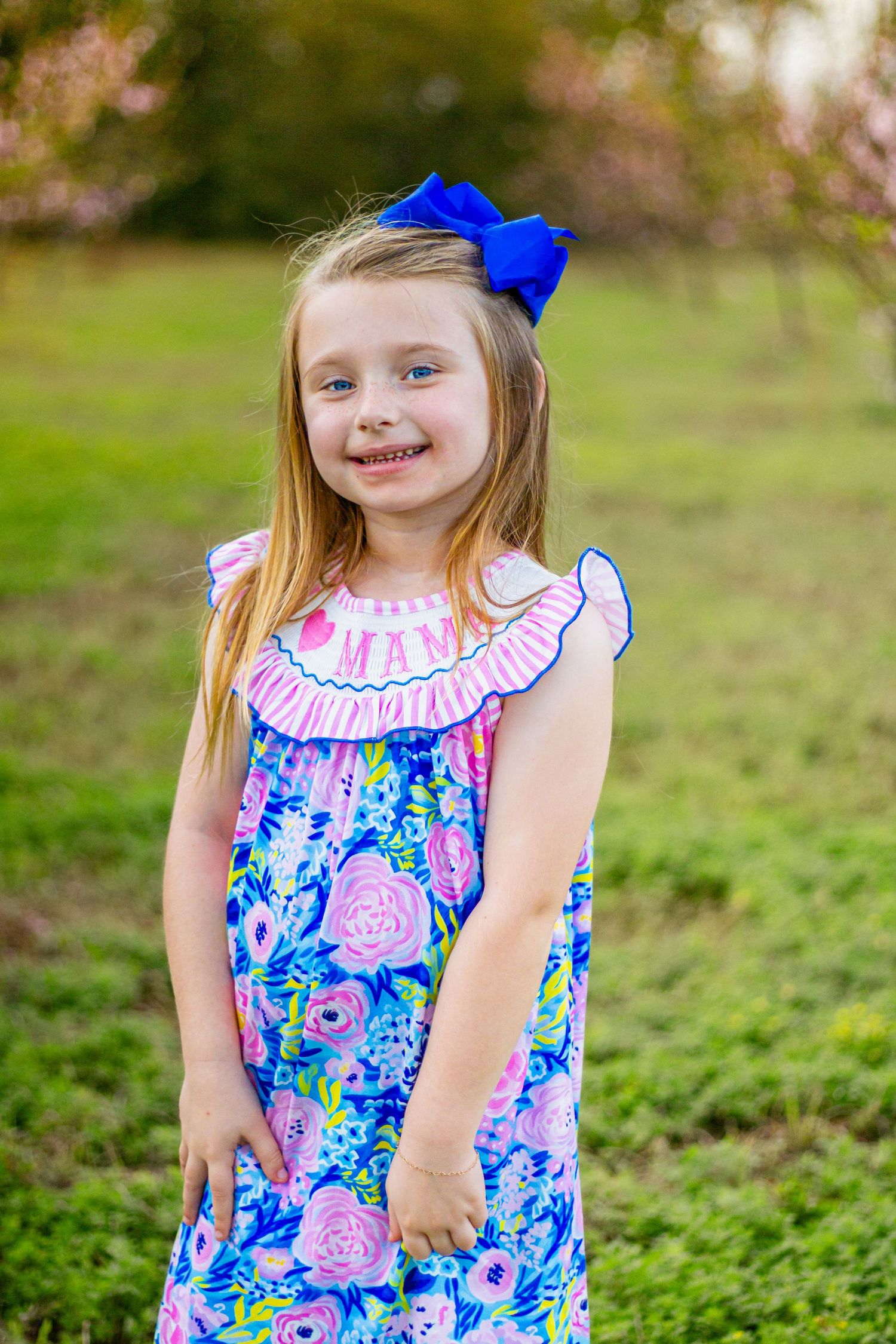 Cheerful blonde girl in vibrant floral dress with blue hair bow smiles in green outdoor spring setting.