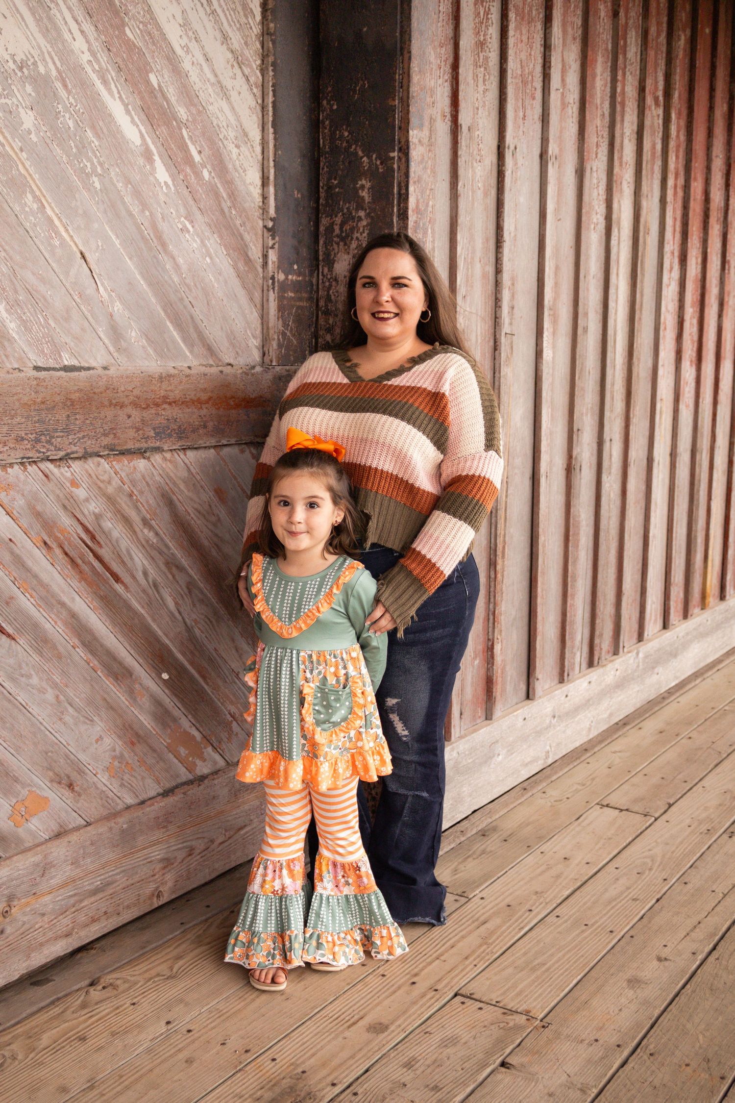A mother and young daughter pose together on a rustic wooden porch in fall outfits.