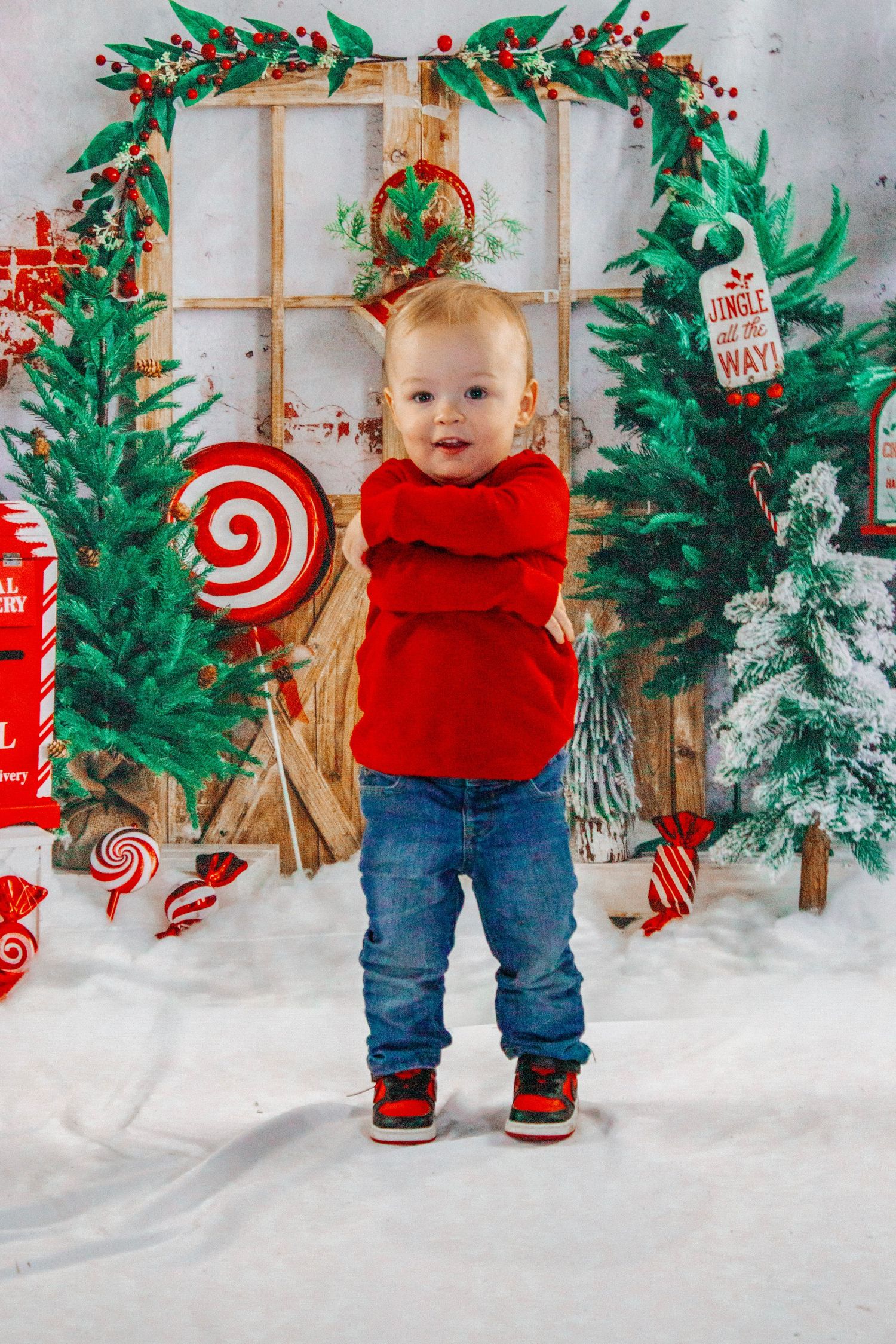 Adorable toddler in red sweater and jeans standing in front of festive Christmas backdrop.