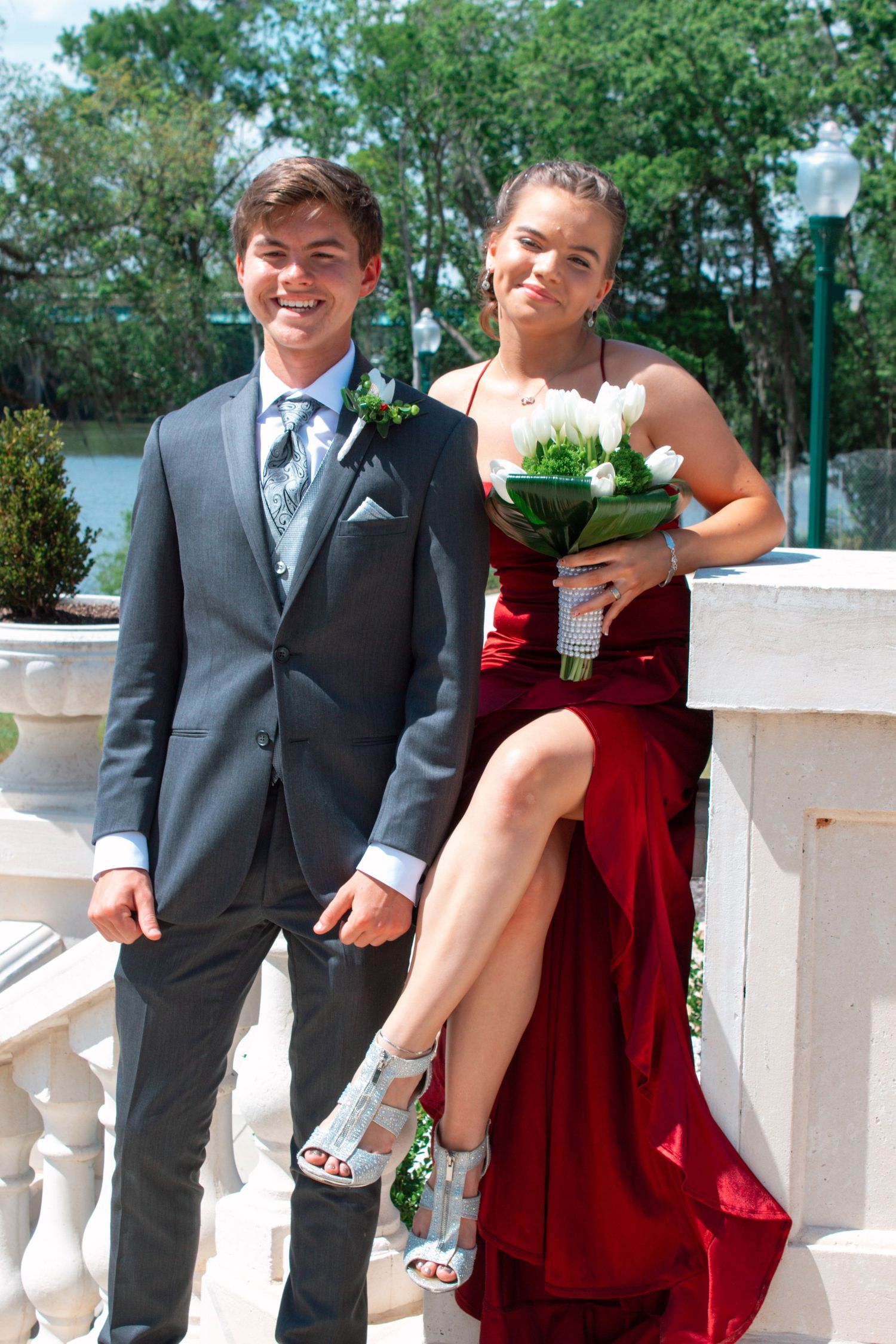 Prom couple poses at white balustrade, girl in red slit gown holding white bouquet, guy in gray suit.