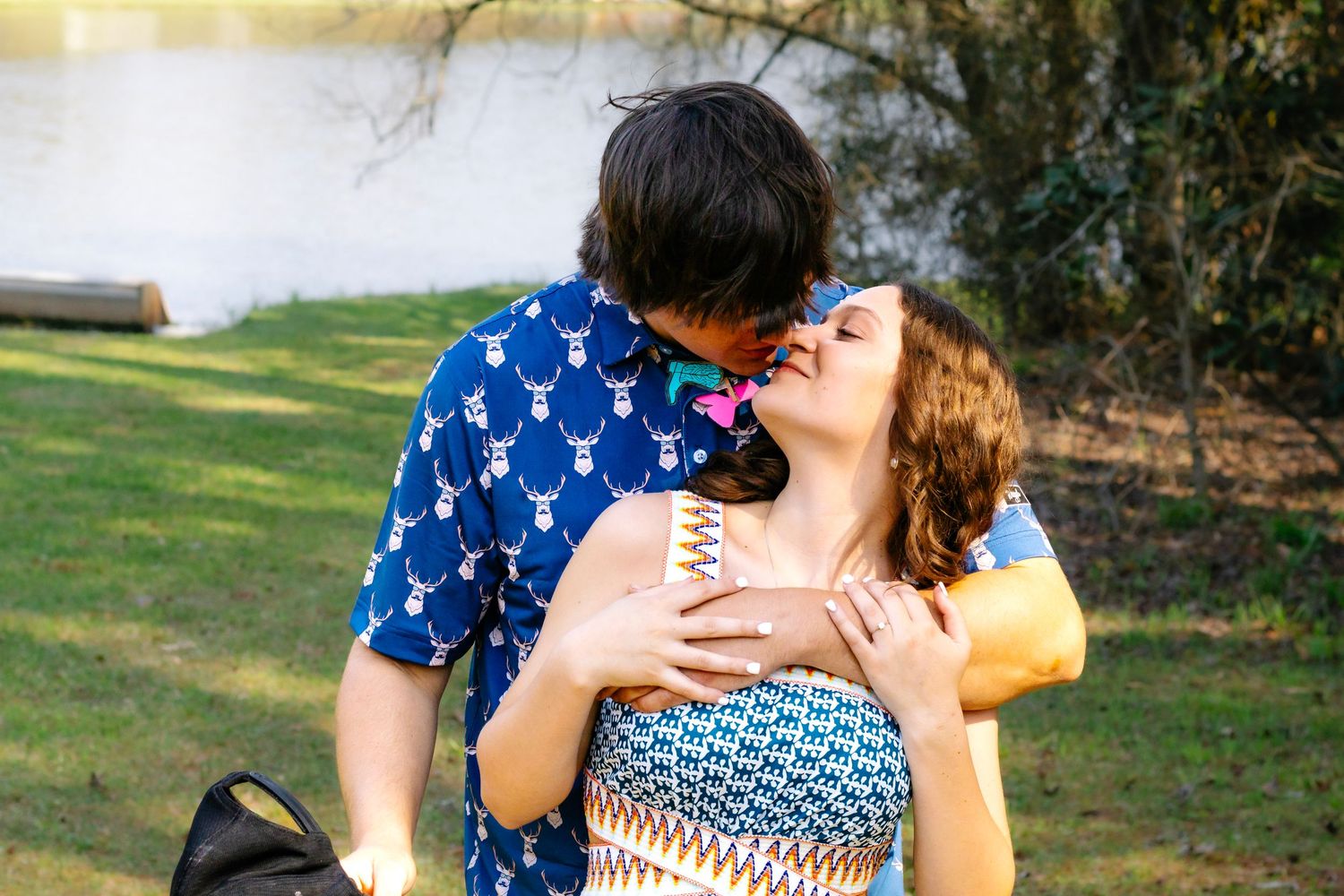 Happy couple laughing and embracing near lakeside, woman in blue patterned dress, man in deer print shirt.