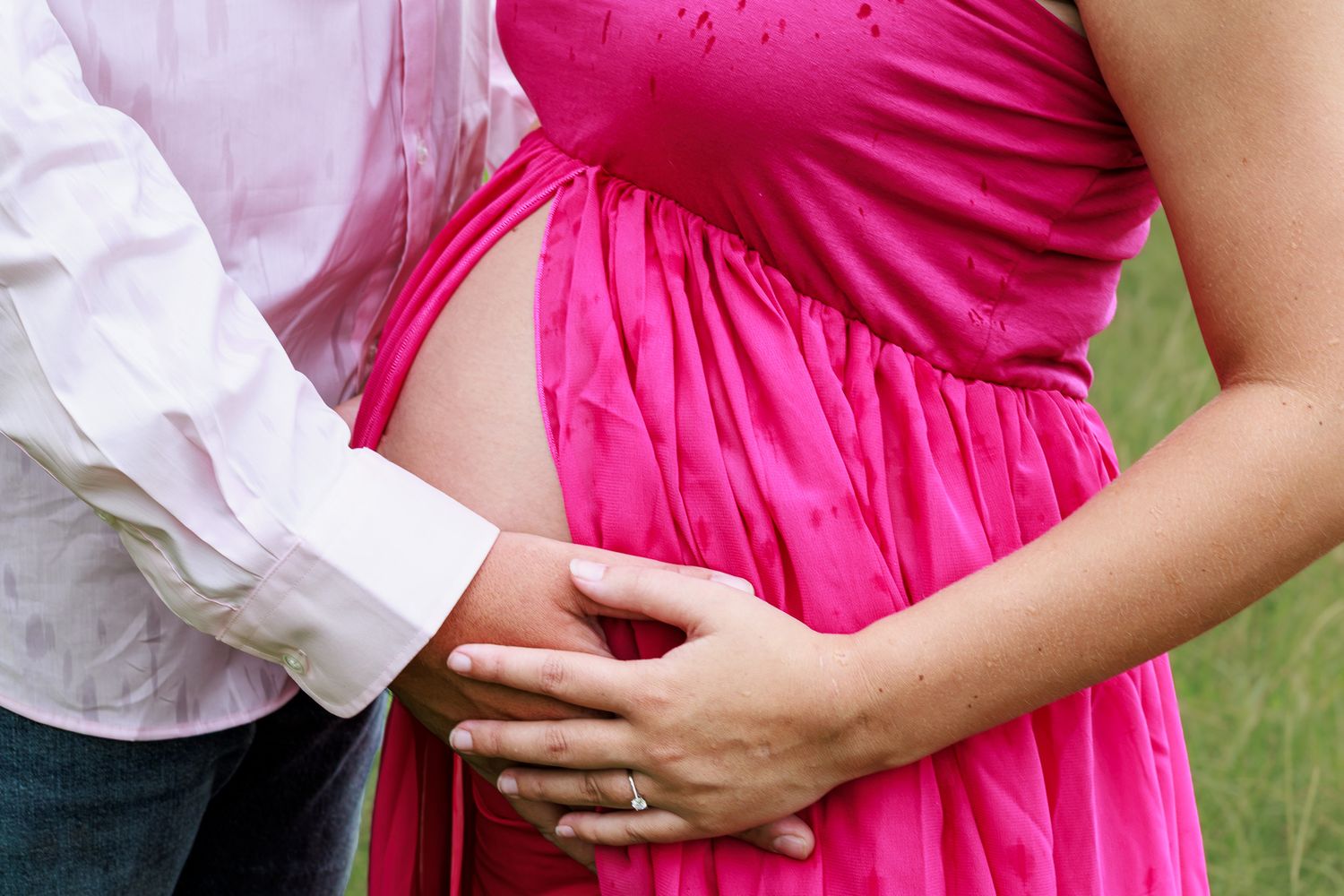 Close-up of pregnant woman in pink dress with partner's hand on baby bump during maternity photoshoot.