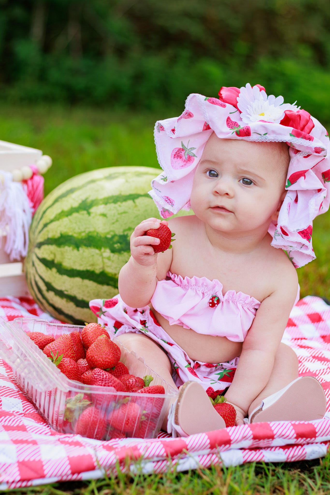 Baby girl in strawberry hat holds a strawberry at a picnic with watermelon and fresh strawberries on a red checkered blanket.
