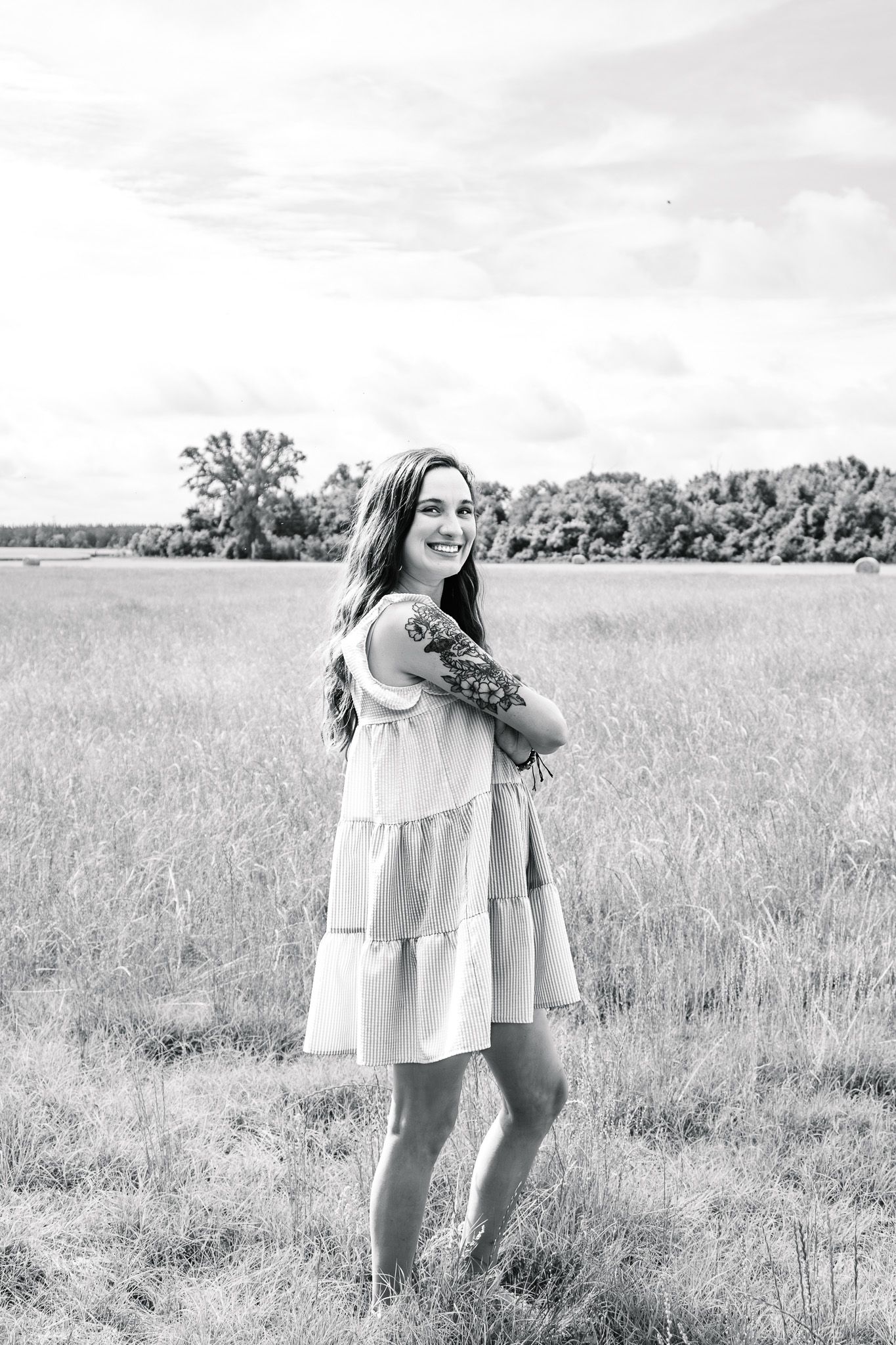 Black and white portrait of a smiling woman in a sundress standing in a field with a floral arm tattoo.