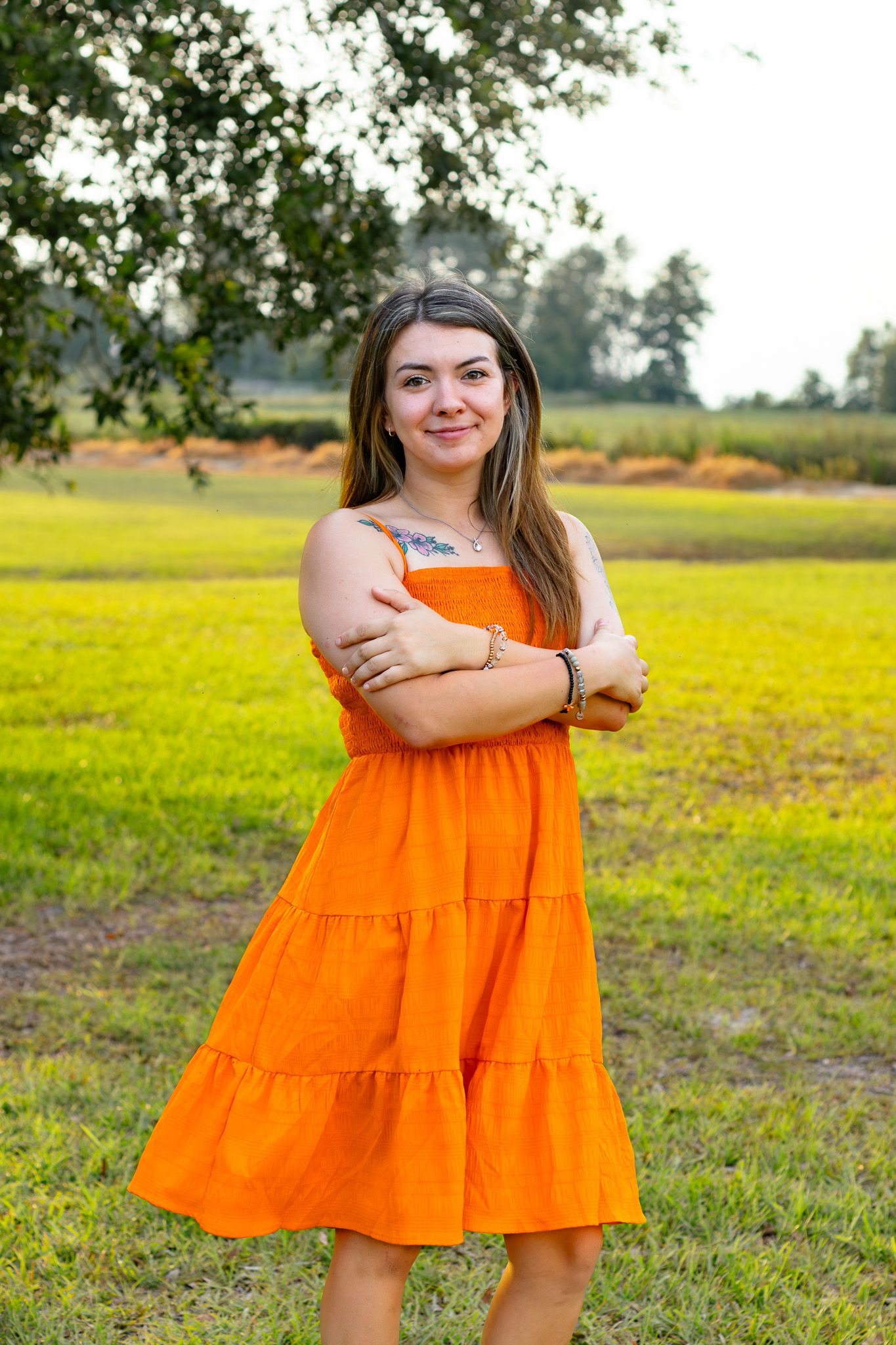 Young woman in orange sundress standing with arms crossed in a sunny green field outdoors.
