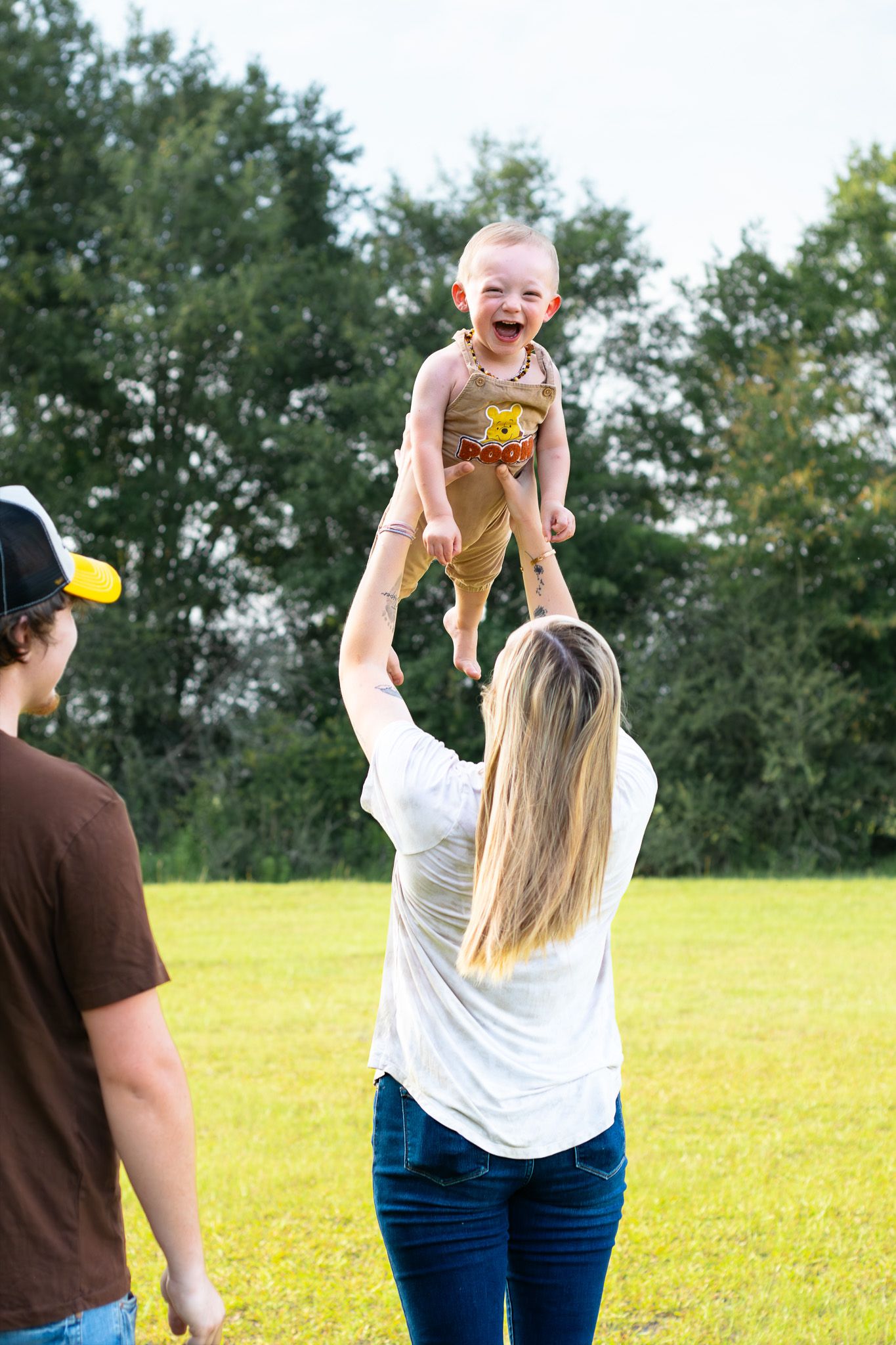 A mother joyfully lifts her laughing baby into the air outdoors while a man watches in a sunny green park.