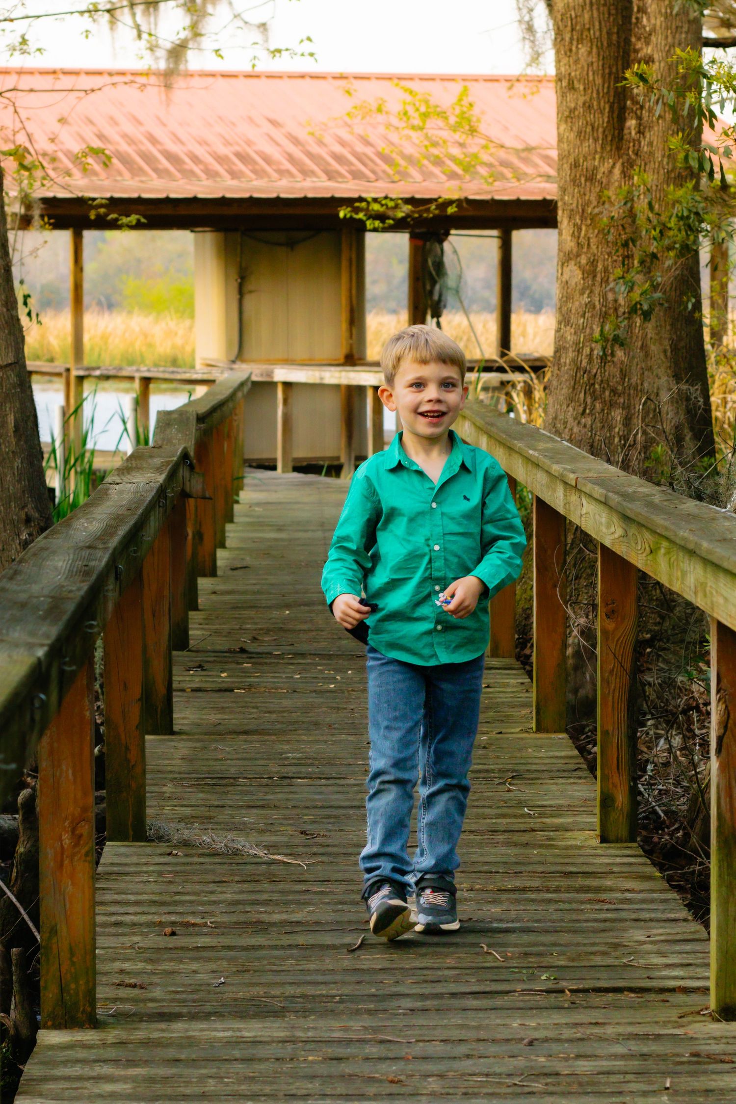 A cheerful young boy in a green shirt and jeans runs along a wooden boardwalk near a rustic building.