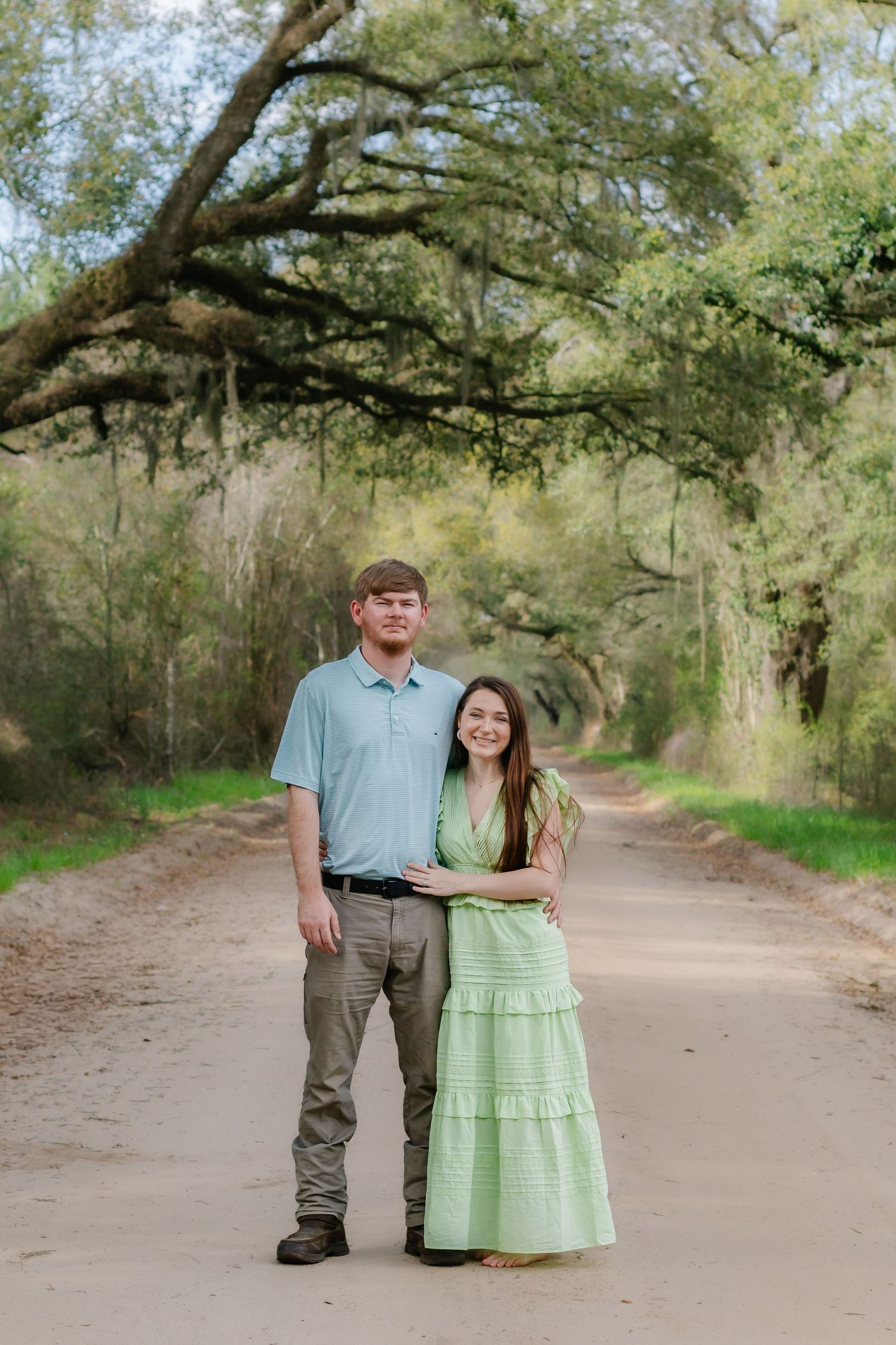 Smiling couple standing together on a scenic tree-lined dirt road, woman in green dress.