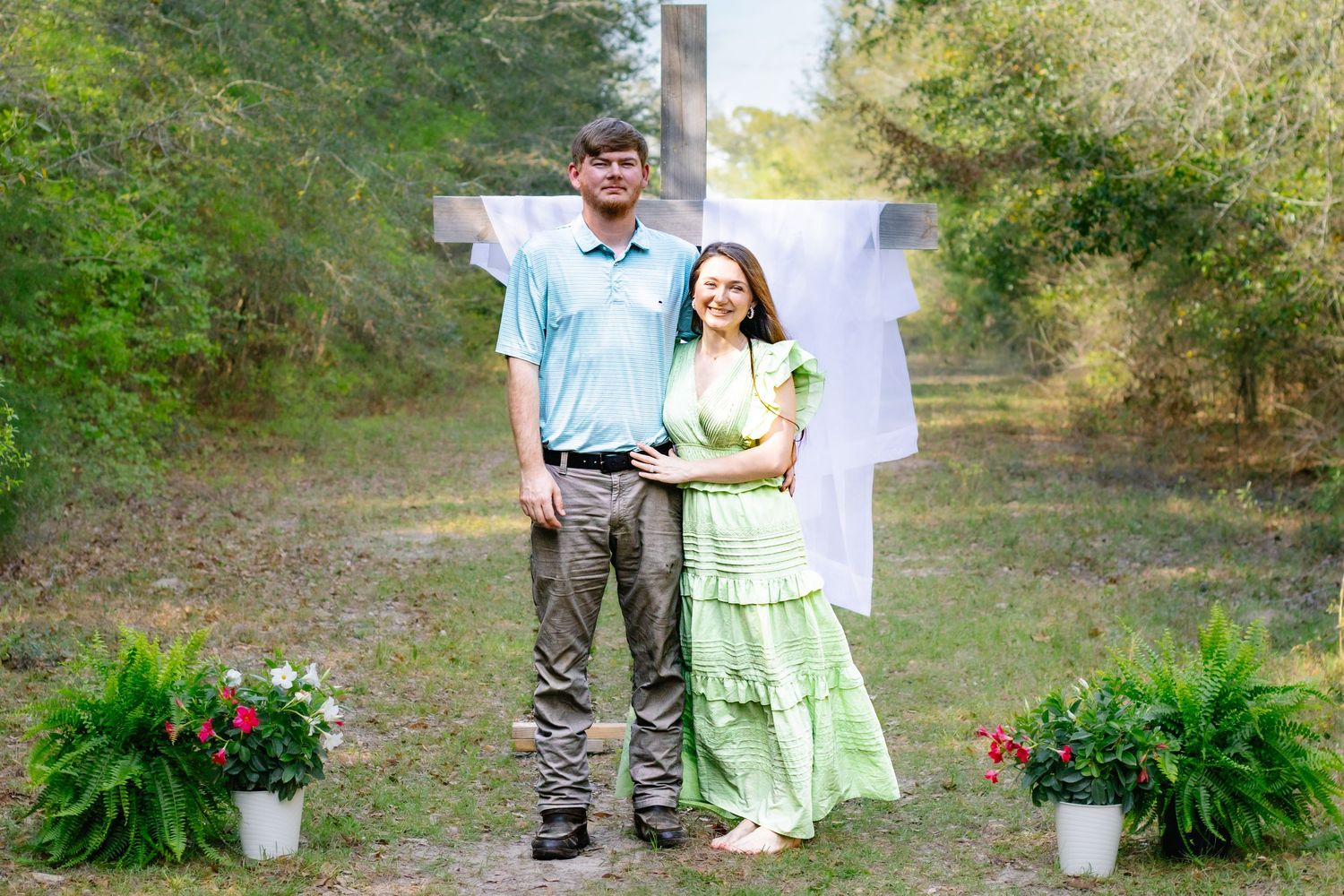 Couple standing before large wooden cross with white drape, surrounded by potted flowers in outdoor setting.