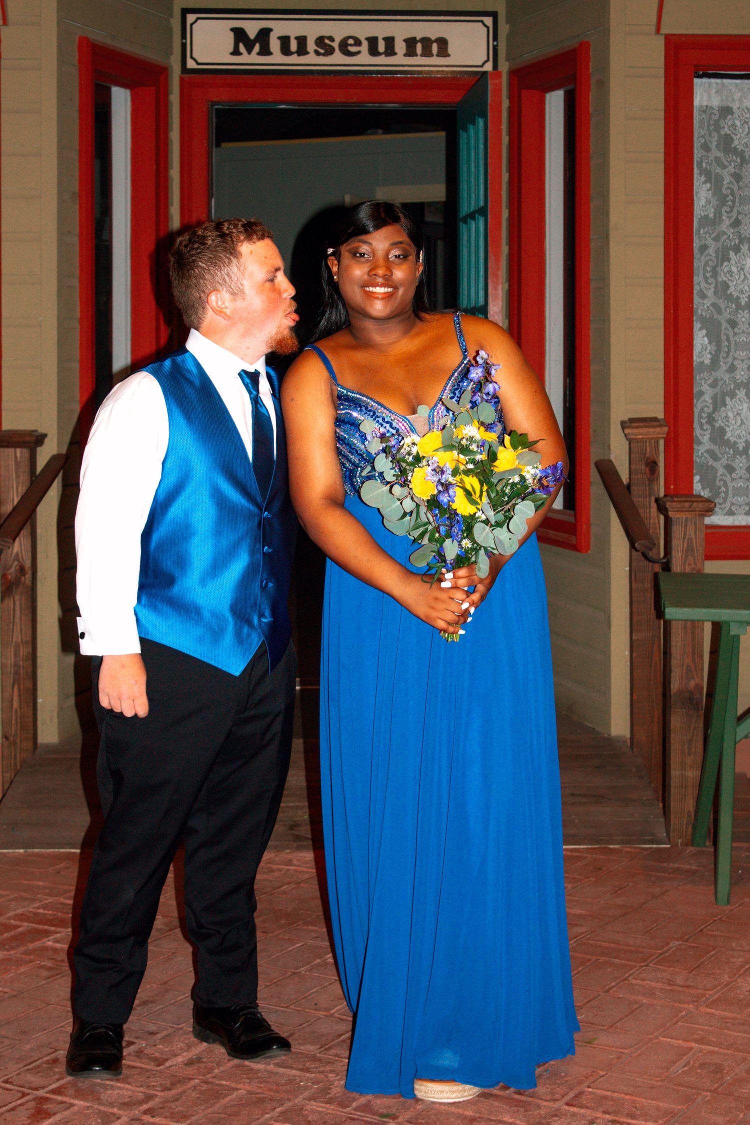A couple in blue formal wear poses outside a museum, woman holding yellow and blue bouquet.
