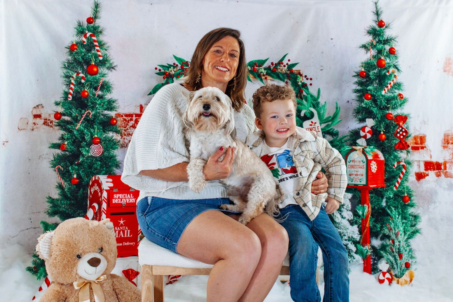 Mother sits with young boy and white fluffy dog in front of Christmas trees during holiday portrait.