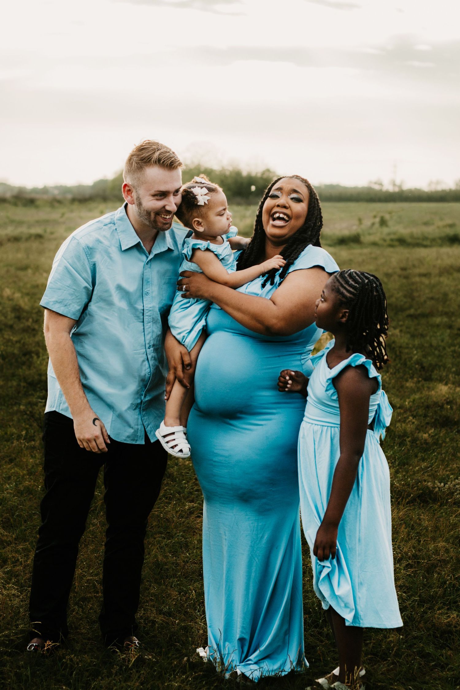 Happy interracial family in matching blue outfits celebrates pregnancy outdoors in green field.