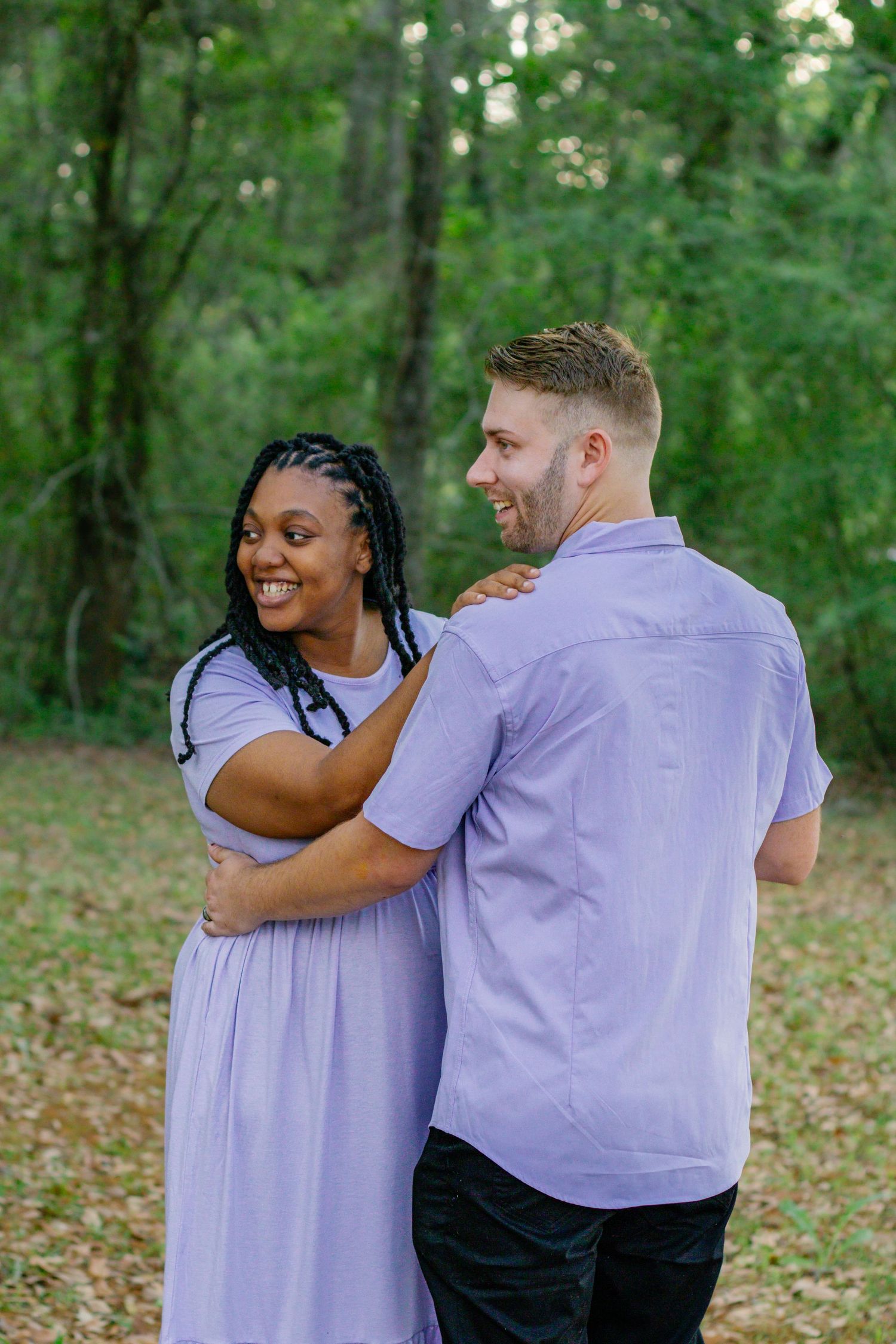 Smiling Black woman in lavender dress embraces man in matching purple shirt outdoors in lush green forest setting.