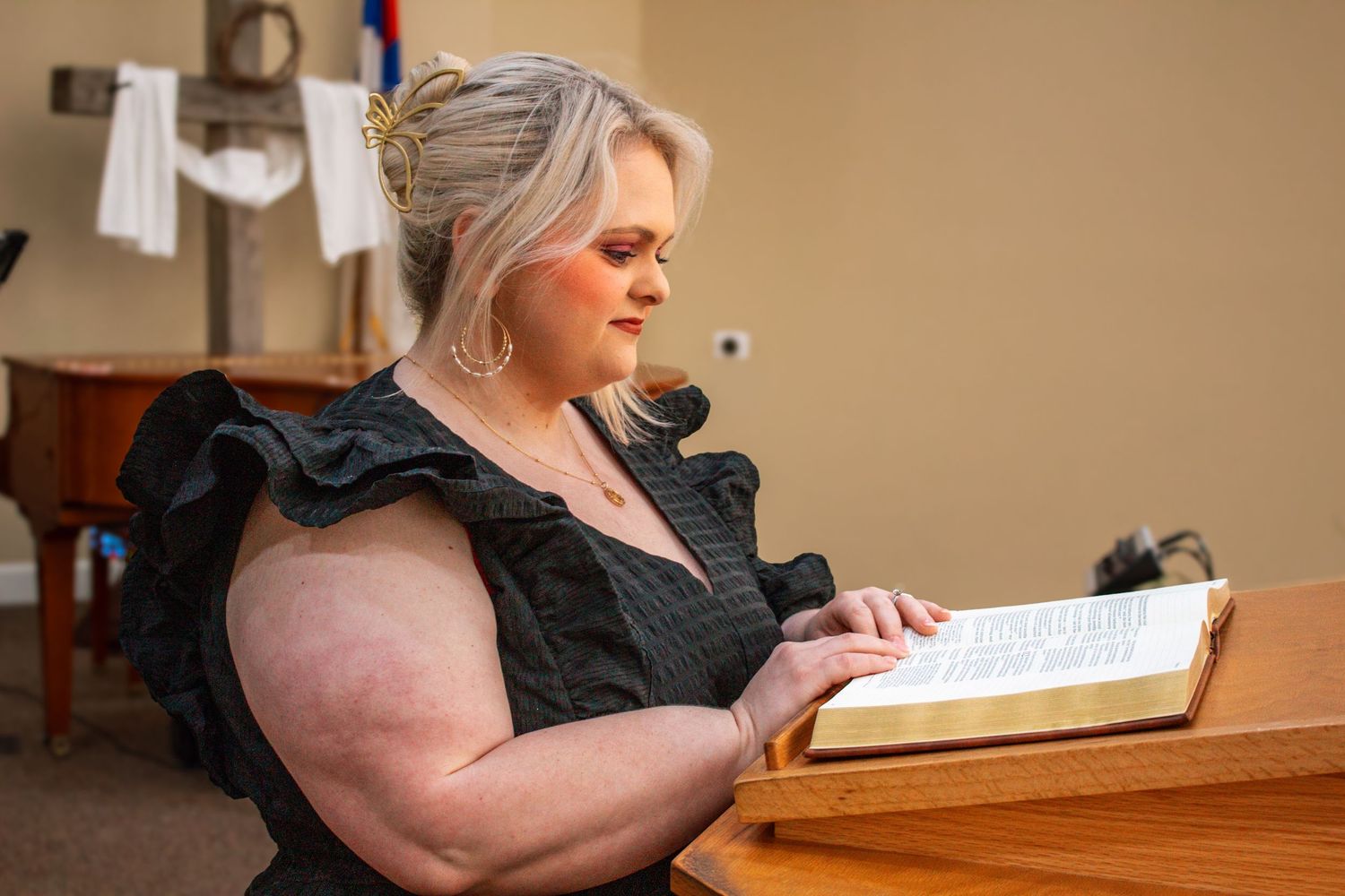 Blonde woman in black dress reading open Bible at wooden church lectern.