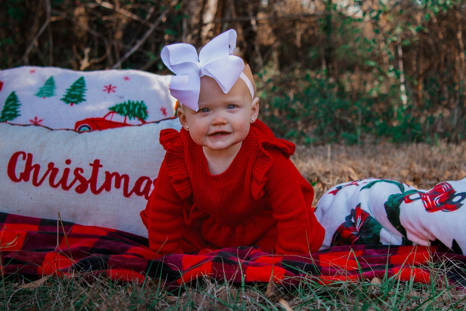Smiling baby girl in red dress with white bow sitting on Christmas blankets outdoors in natural setting.