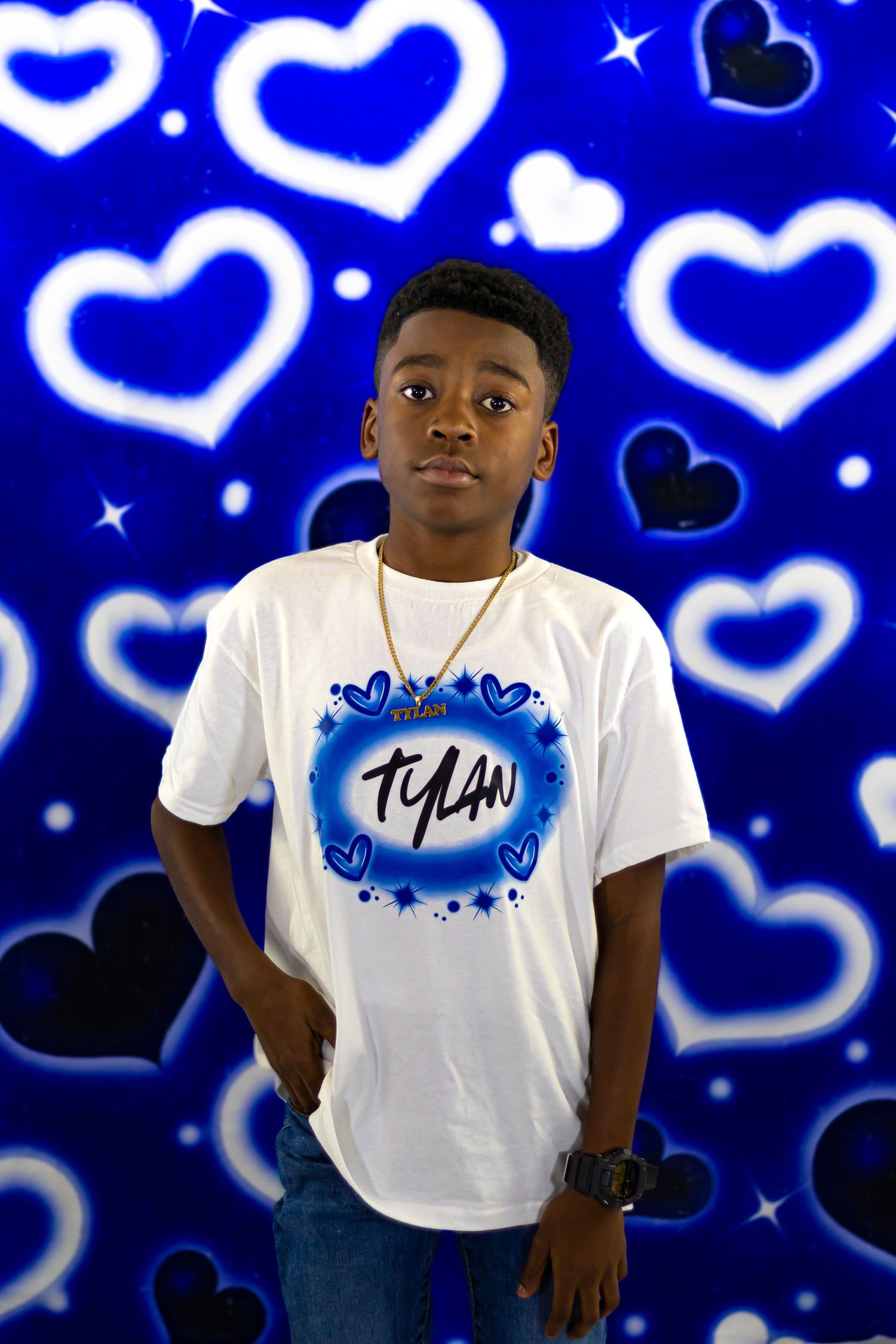 Young Black boy named Tylan in custom airbrush tee poses against glowing blue hearts backdrop.