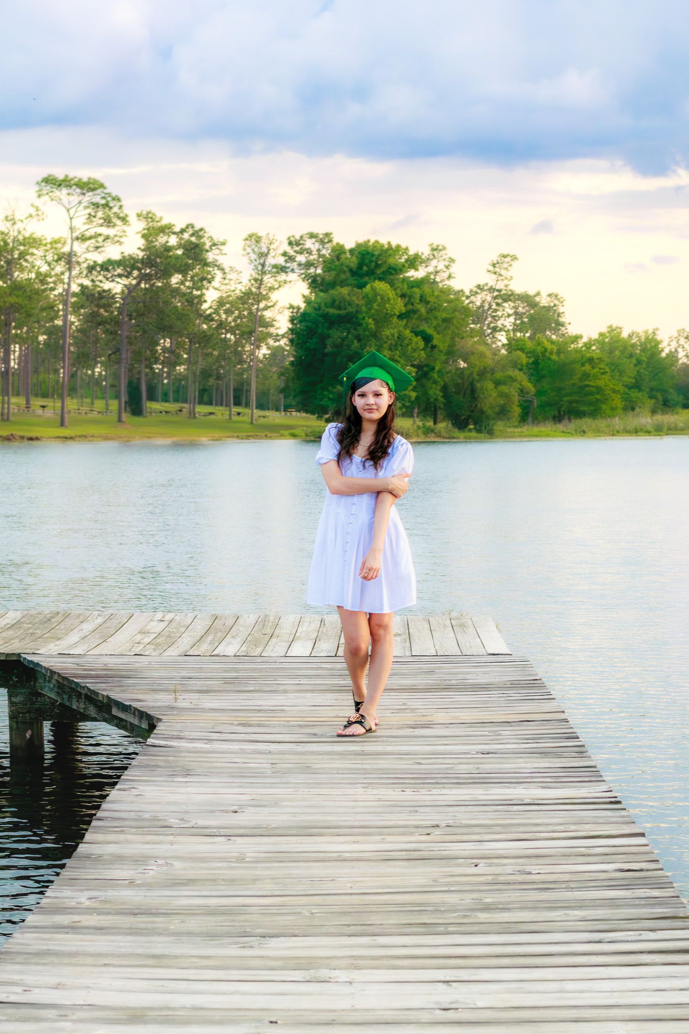 Young woman in white dress and green graduation cap walks on wooden dock by a serene lake.