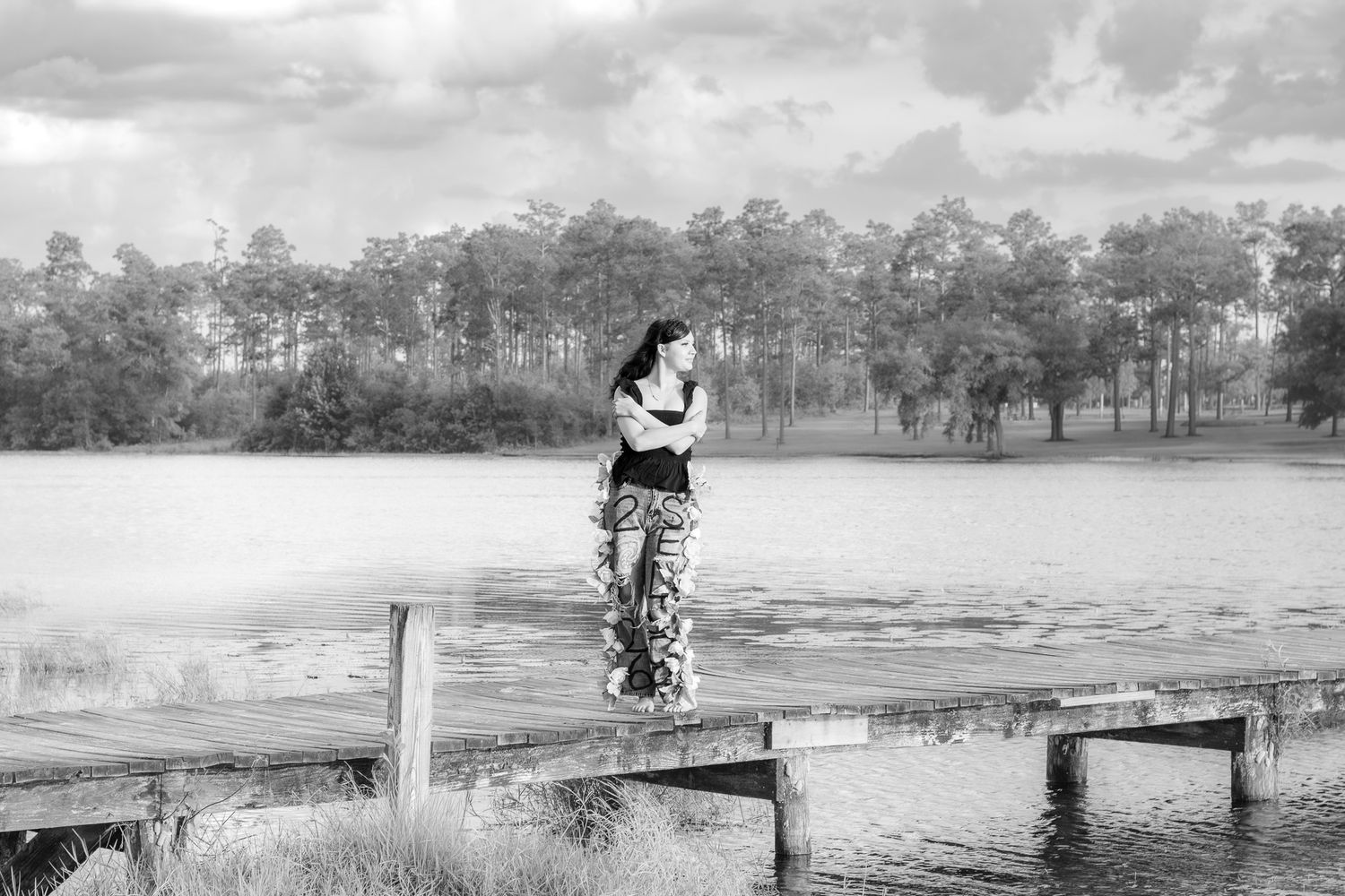 Black and white photo of a woman standing on a wooden dock by a serene lake surrounded by trees.