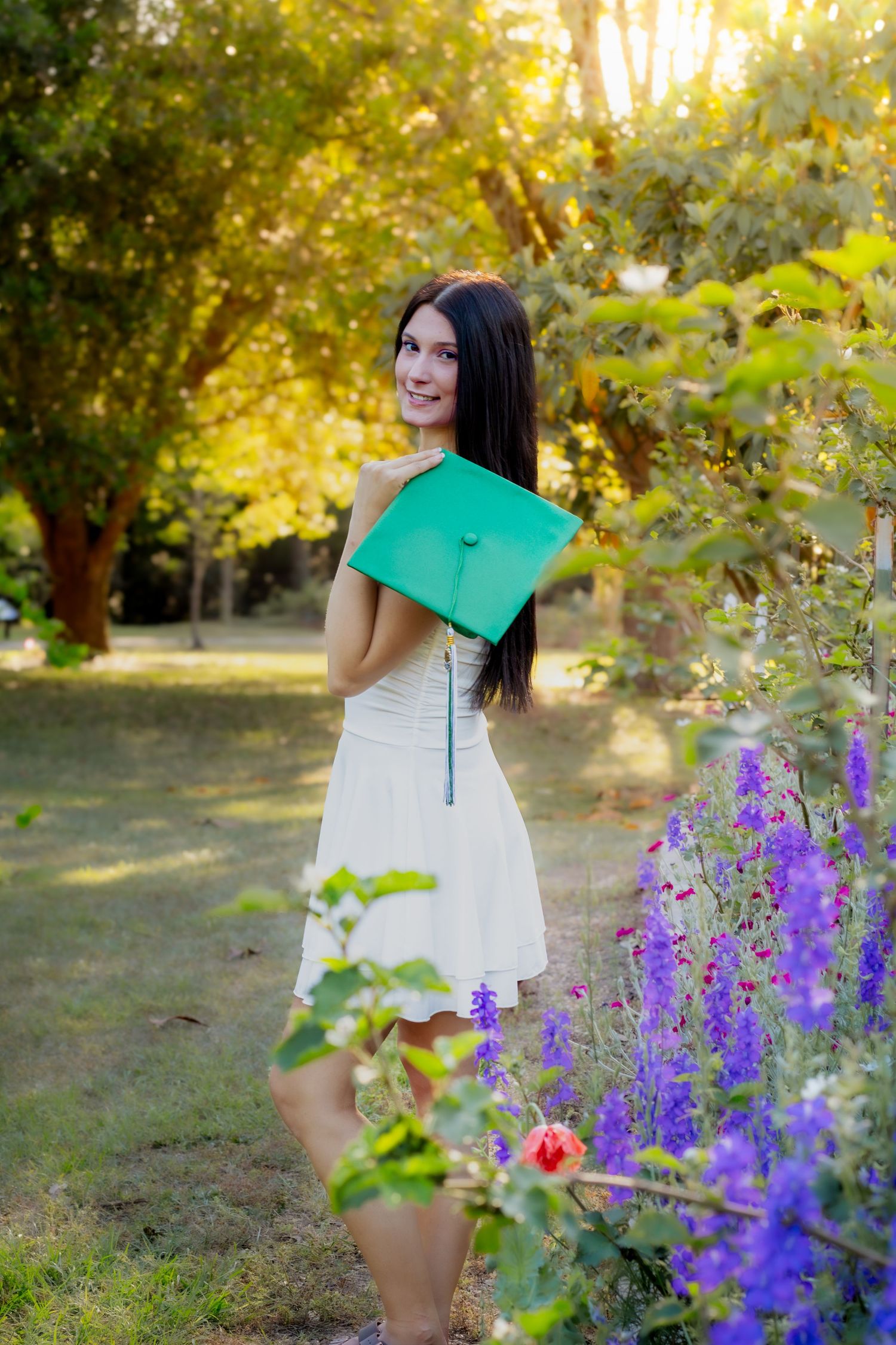 Happy young woman in white dress holds green graduation cap among colorful garden flowers at sunset.