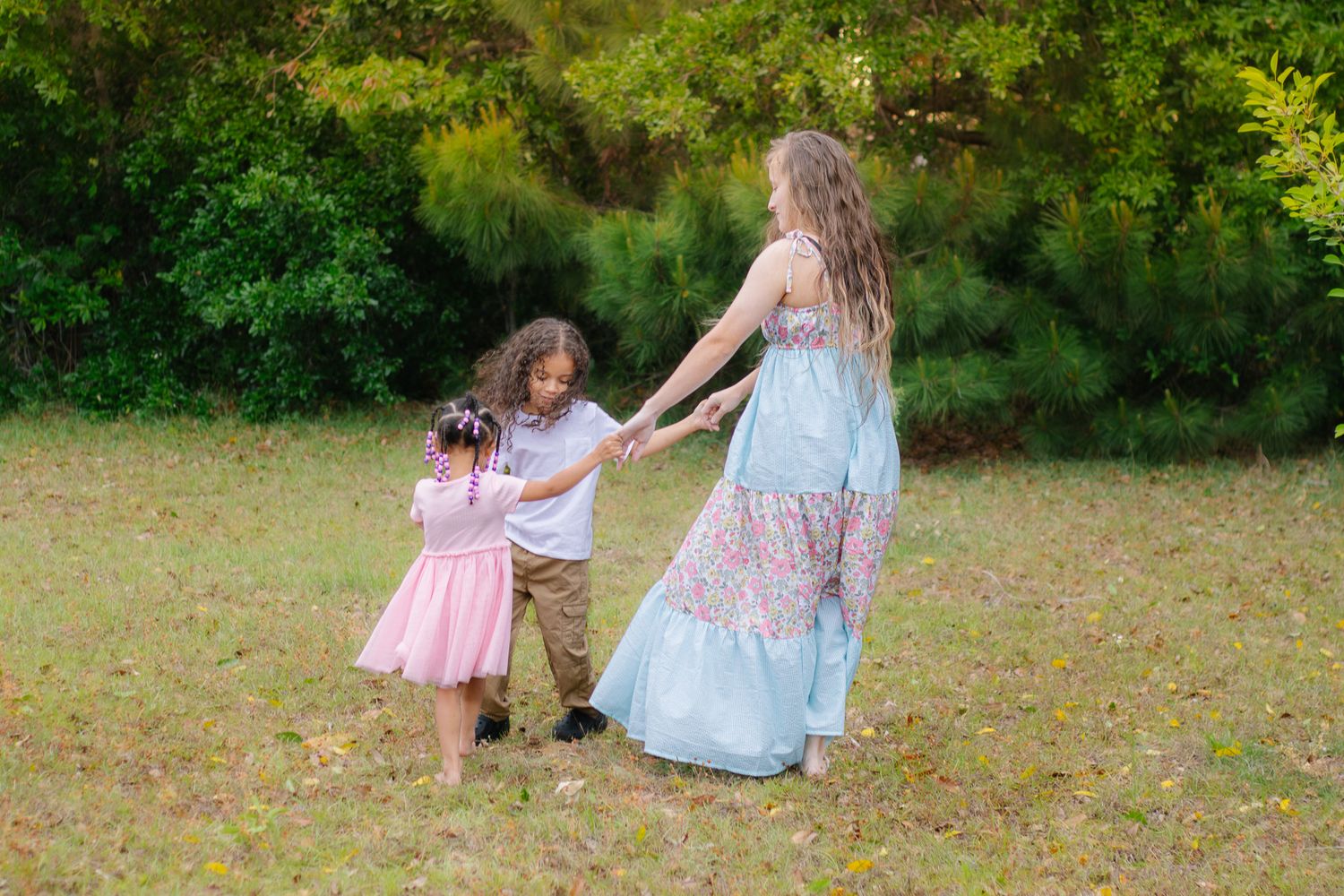 Woman in blue floral maxi dress plays with two young children in a grassy outdoor setting with trees.