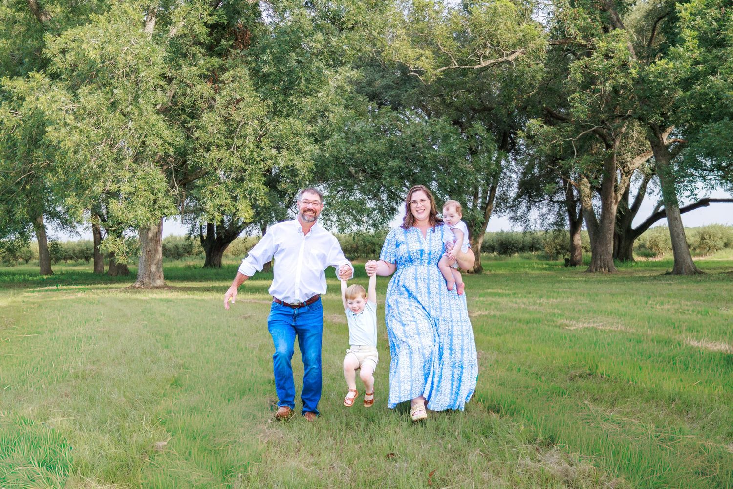 Family of four walking through a lush green park, parents holding toddler's hands, mother carrying baby in blue tie-dye dress.