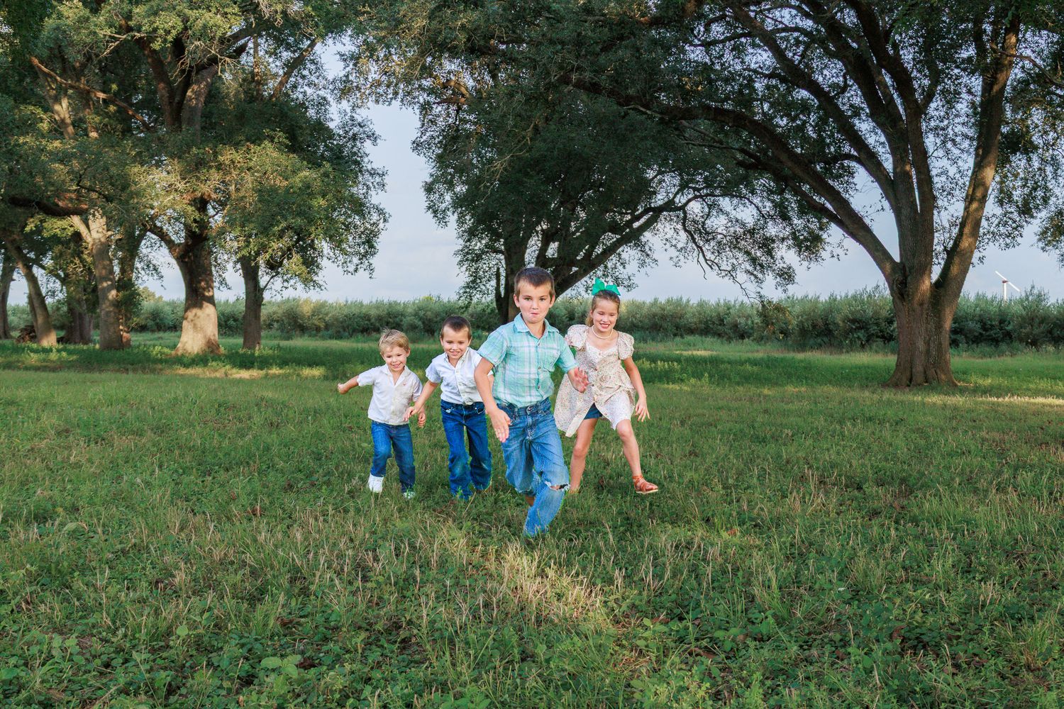 Four young children running and playing together in a lush green park surrounded by tall trees.