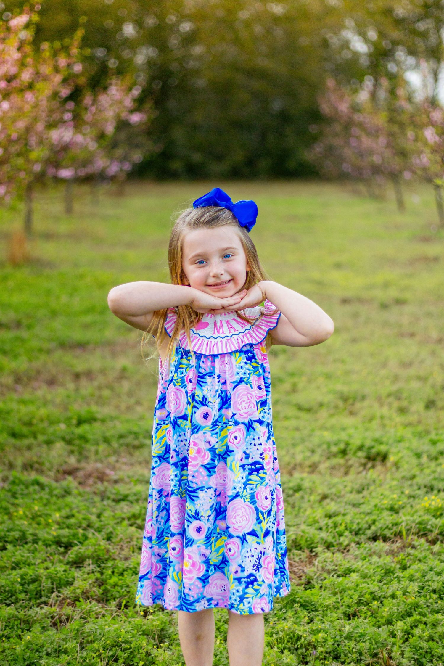 Smiling blonde girl in colorful floral dress with blue bow poses hands under chin in spring garden setting.