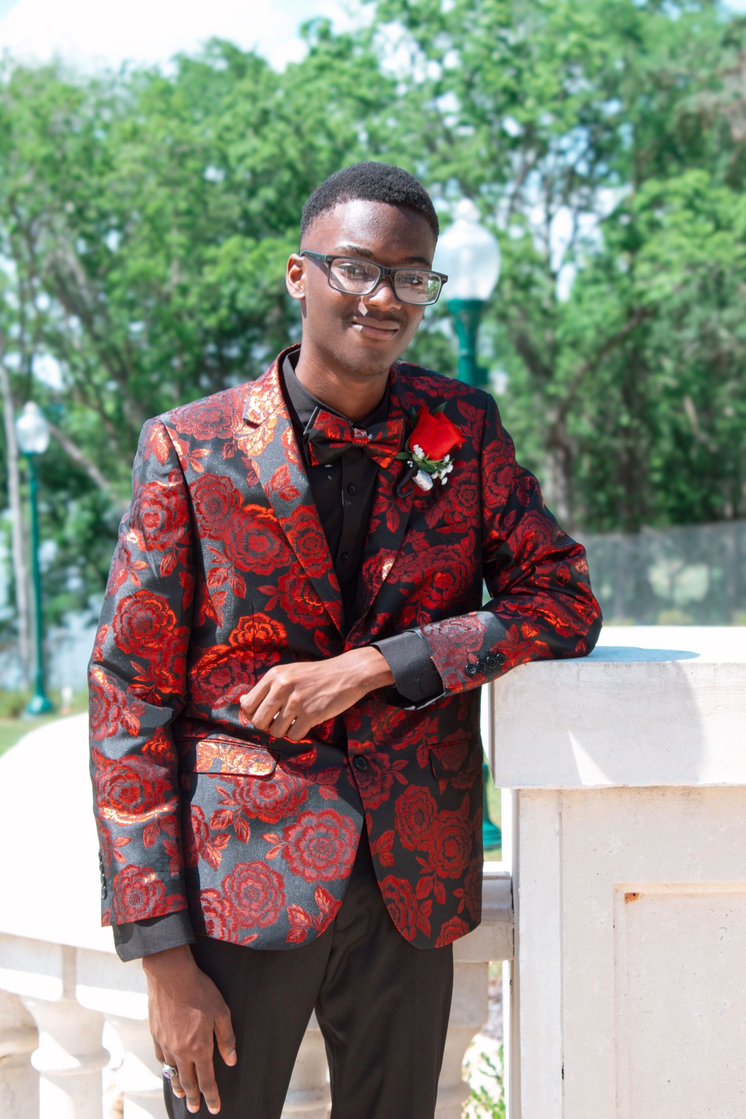 Smiling young man in red floral blazer with boutonniere leans against white railing at prom.
