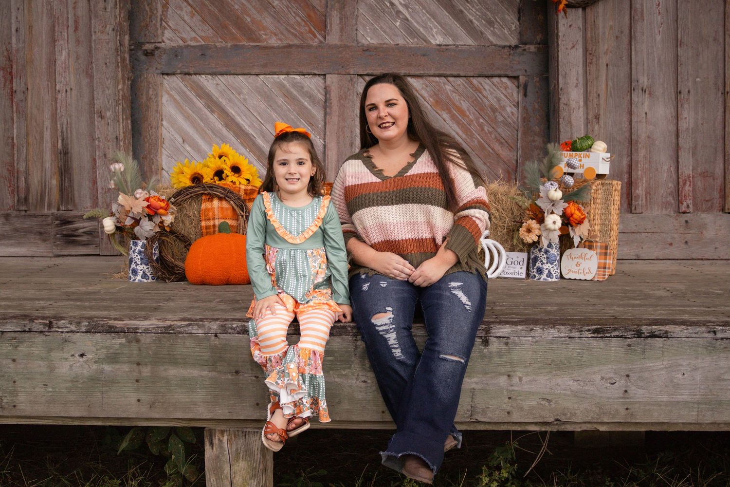A mother and daughter sit together on a wooden bench with fall pumpkin decorations behind them.