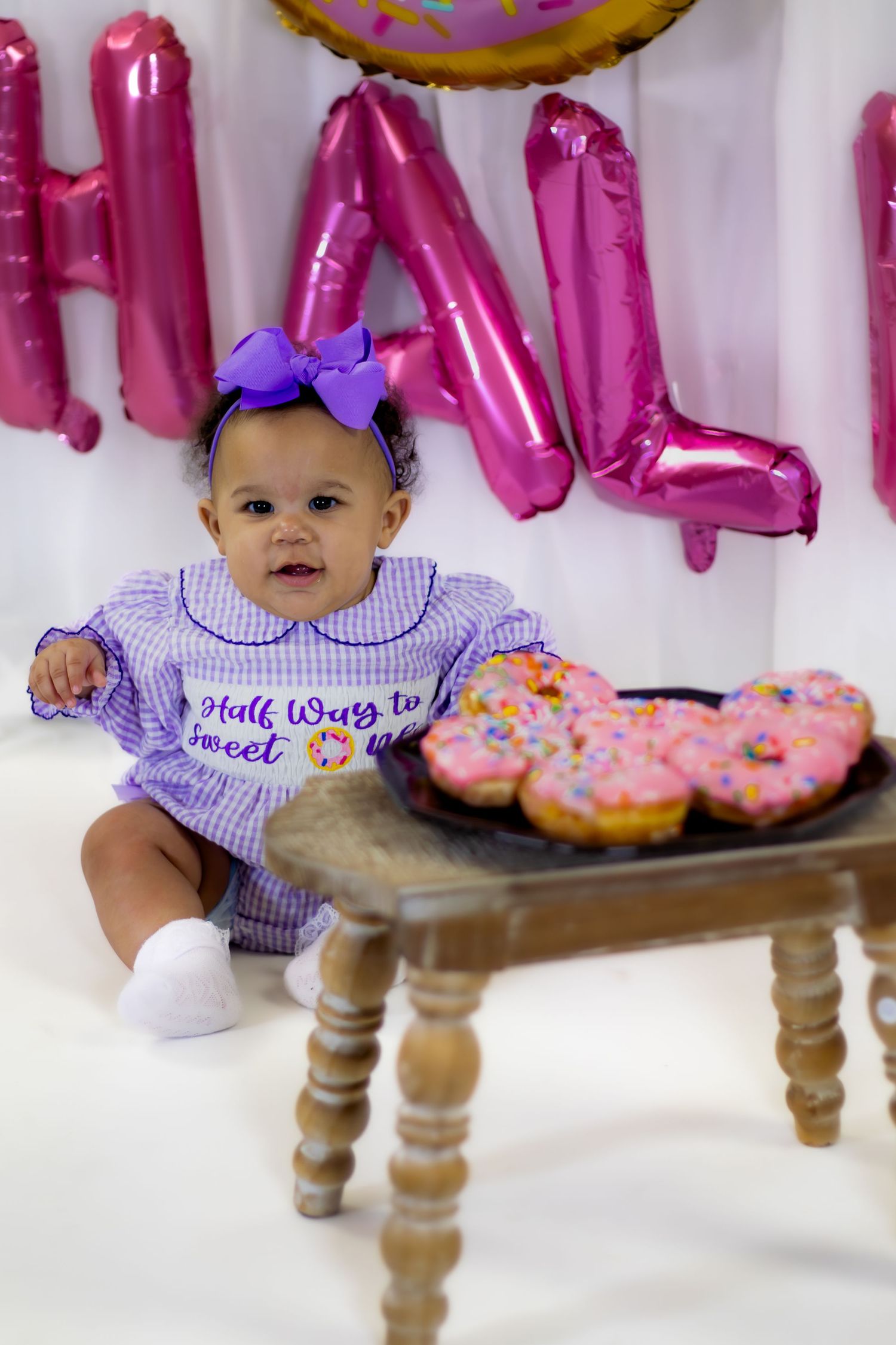 Baby girl in purple gingham romper with bow sits beside pink donuts on wooden stool at half birthday.