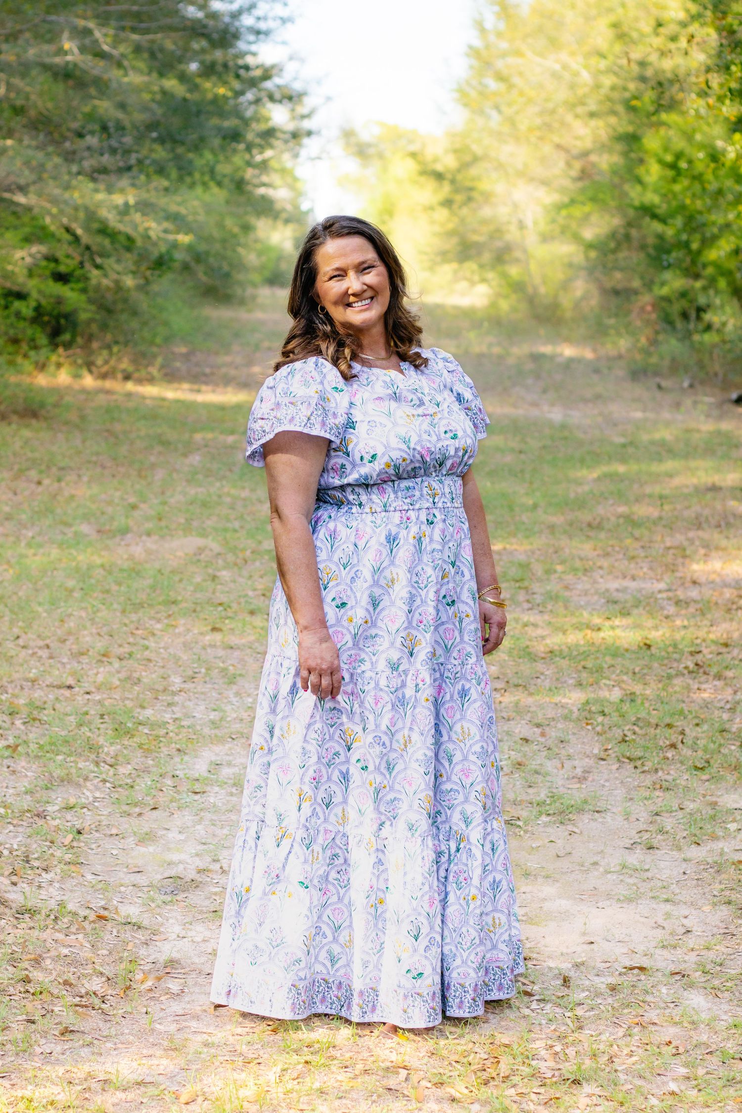 Smiling woman in a blue floral maxi dress standing on a sunlit dirt path in nature.