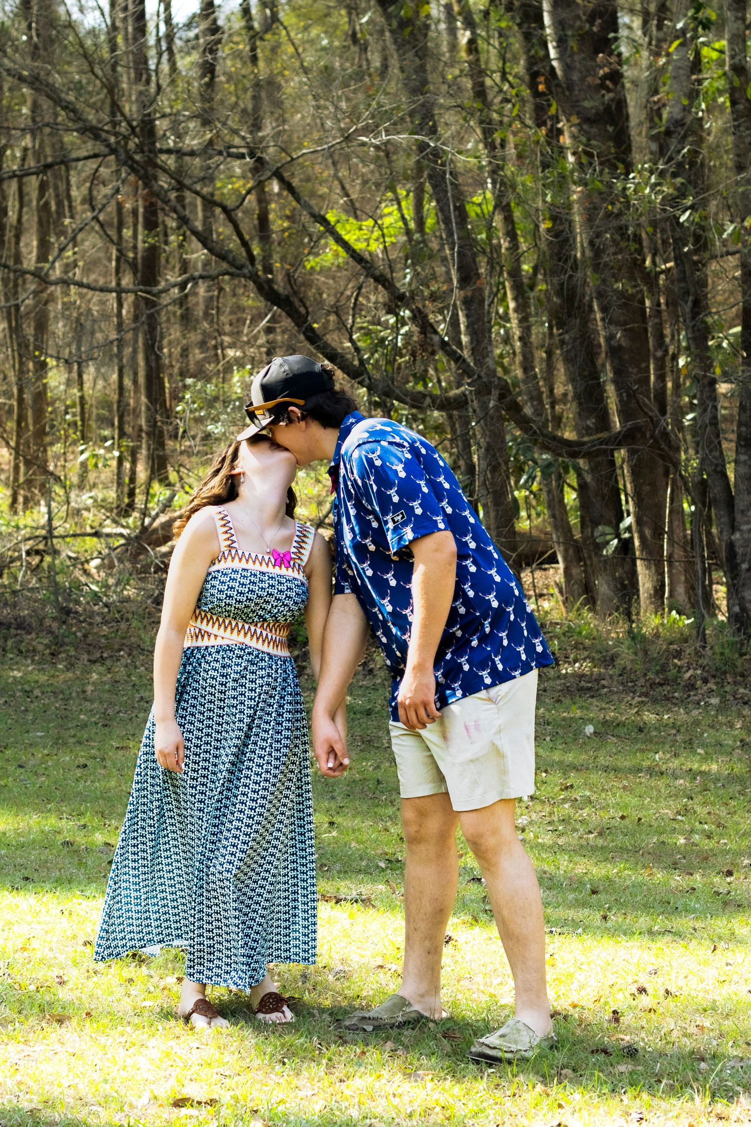Couple sharing a kiss while holding hands in sunlit park, man in blue deer print shirt and khaki shorts.