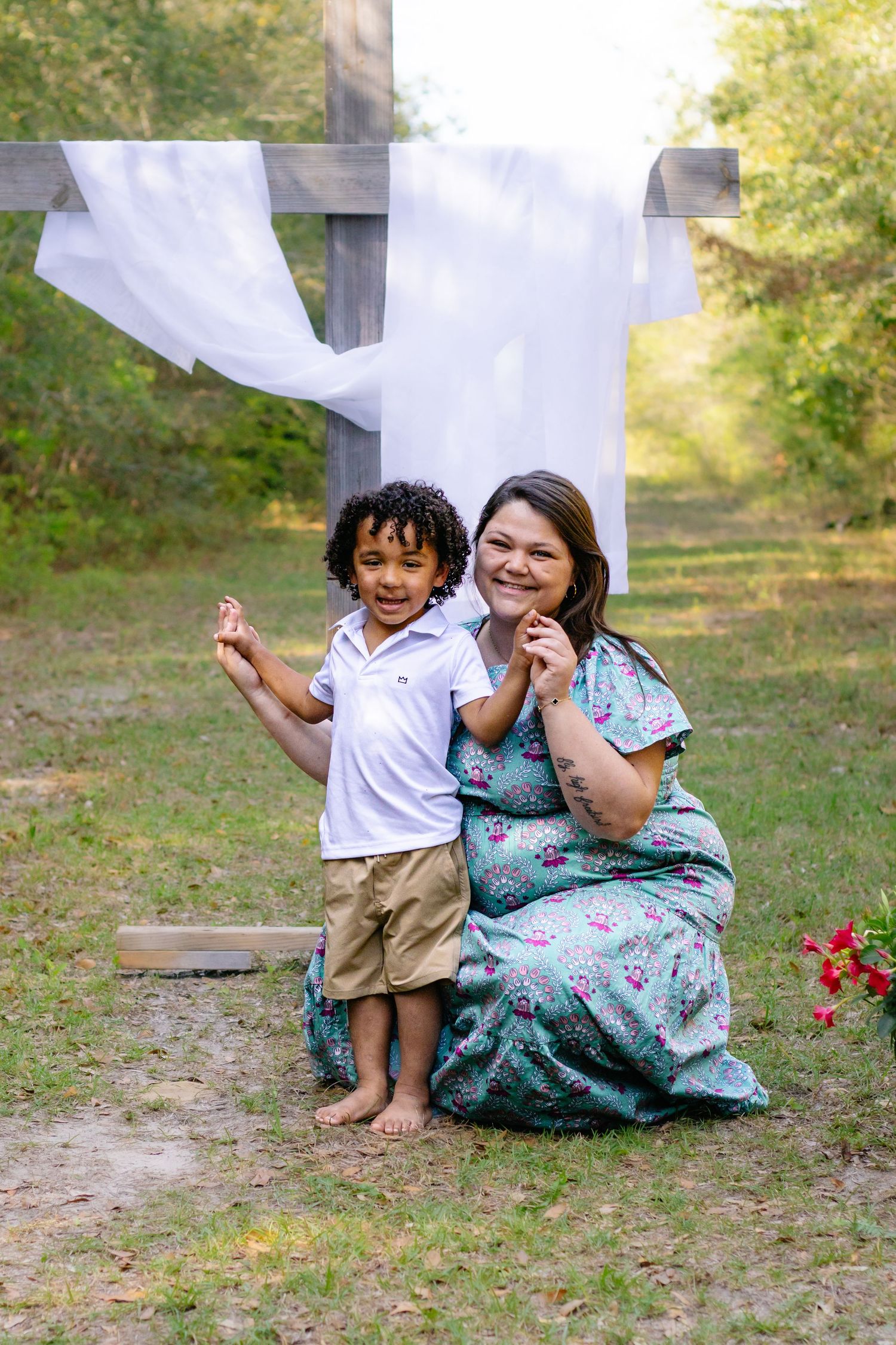 A woman in a floral dress kneels beside a young curly-haired boy in front of a wooden cross outdoors.