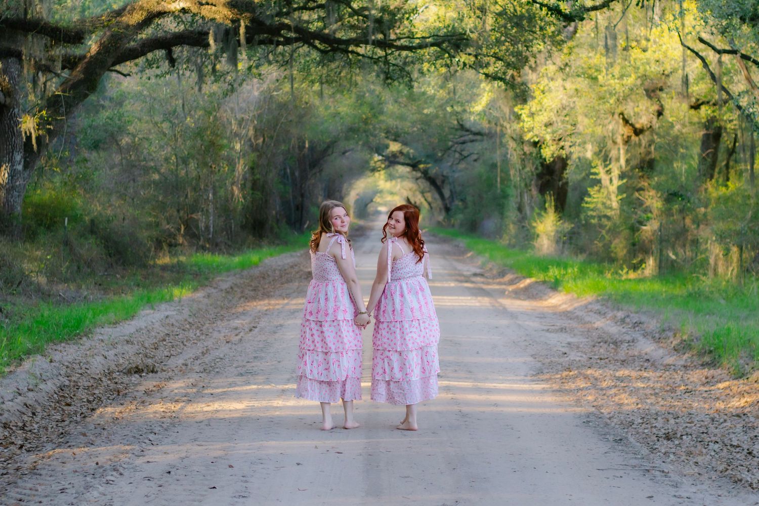 Two girls in pink dresses holding hands on a beautiful tree-lined dirt road at golden hour.