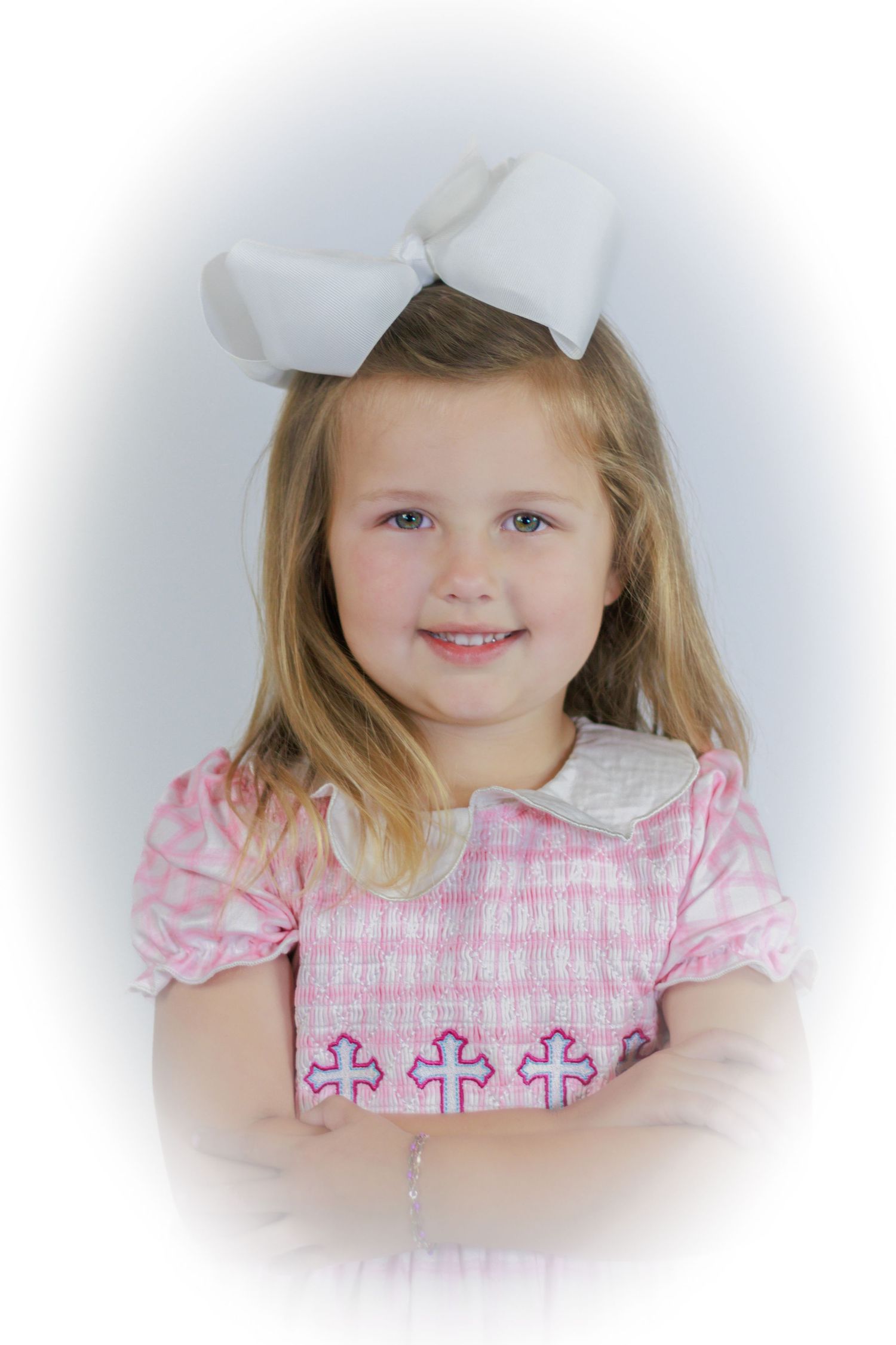 Young girl in pink plaid dress with cross embroidery and white bow hair accessory smiling for a portrait photo.