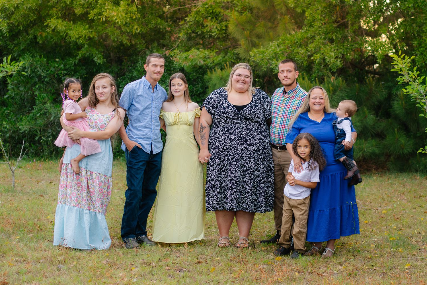 Large family group portrait of eight people posing together outdoors in grassy area with trees.