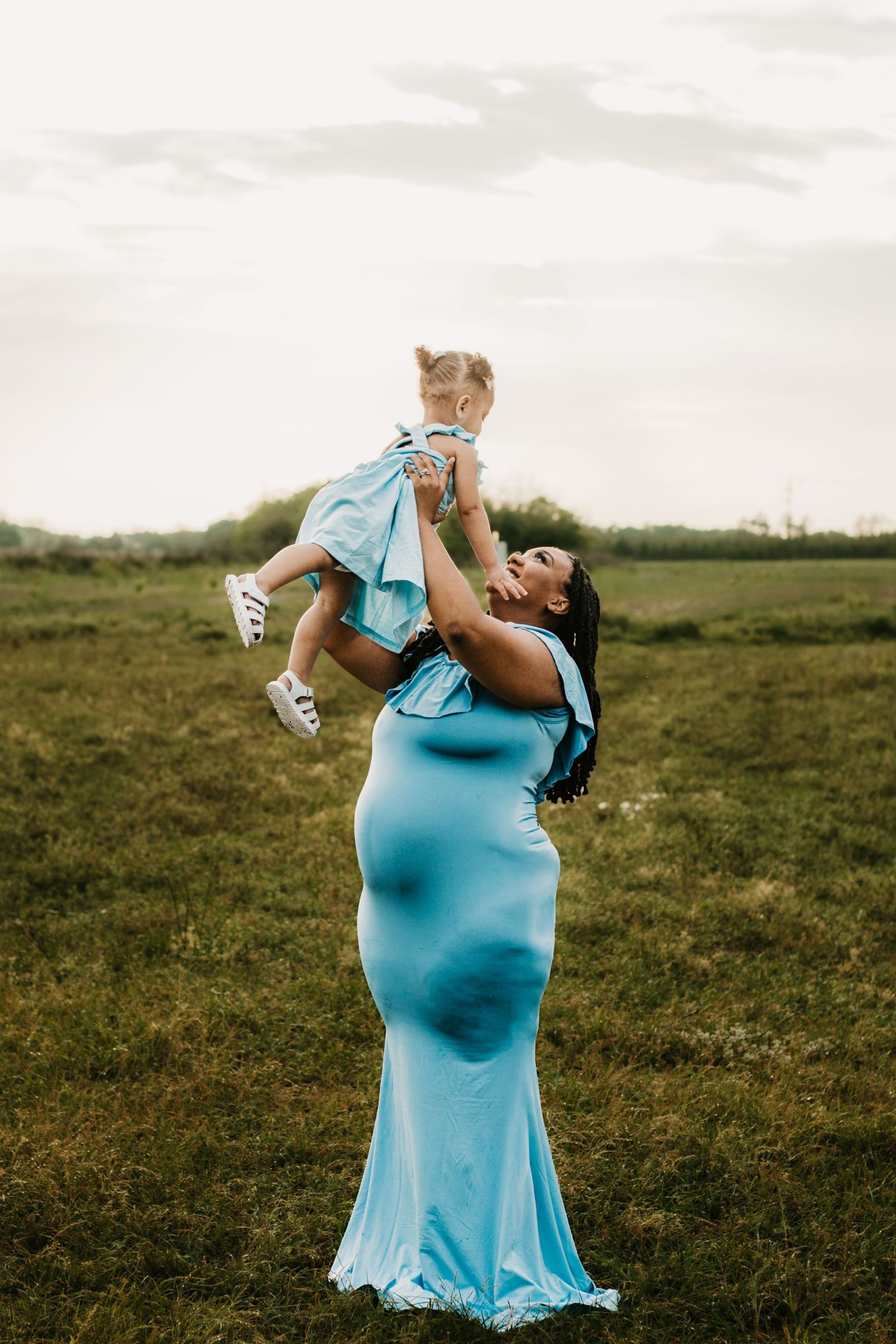 Pregnant woman in blue gown lifting toddler in matching outfit in green field.