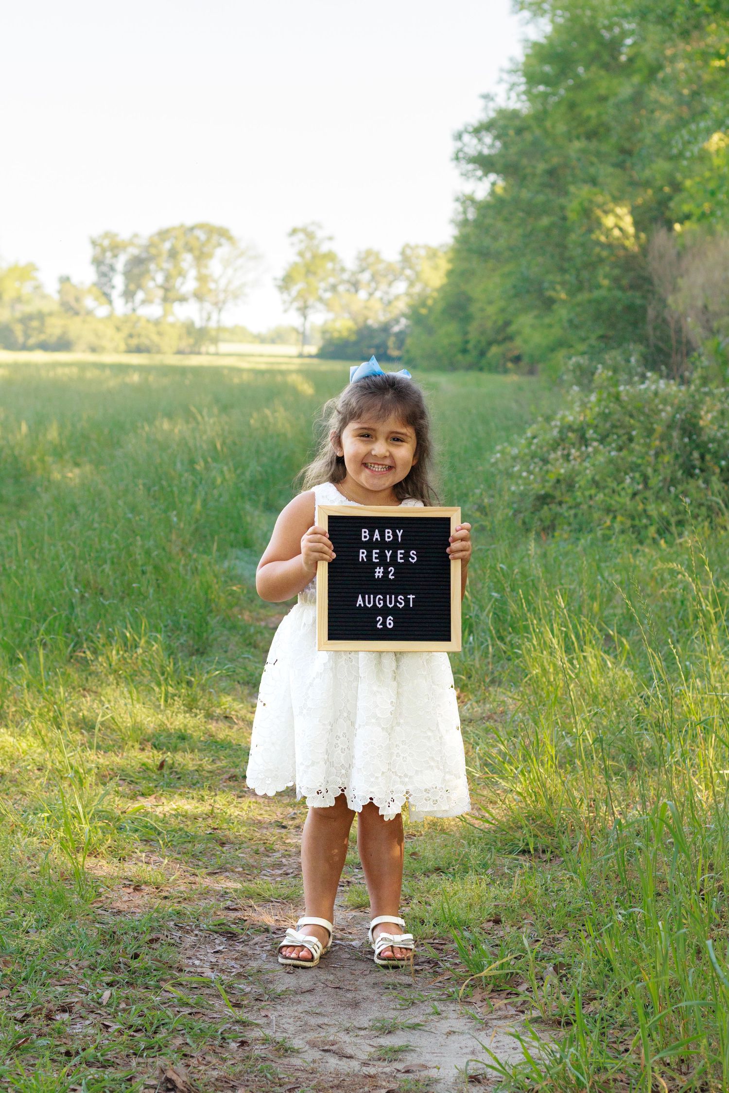 Smiling girl in white dress holds letter board announcing Baby Reyes #2 arriving August 26 outdoors.