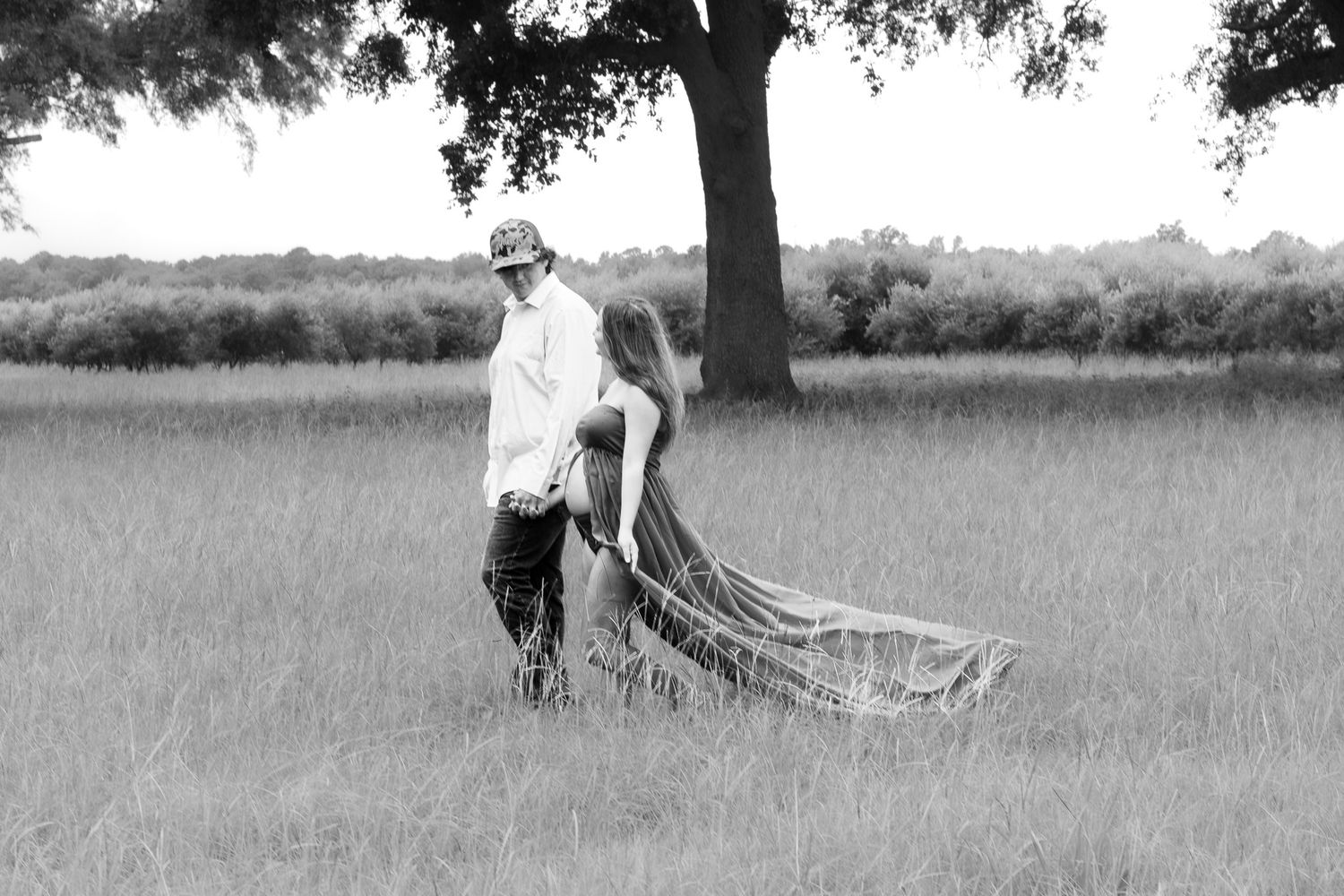 Black and white photo of couple walking in field with flowing dress train.