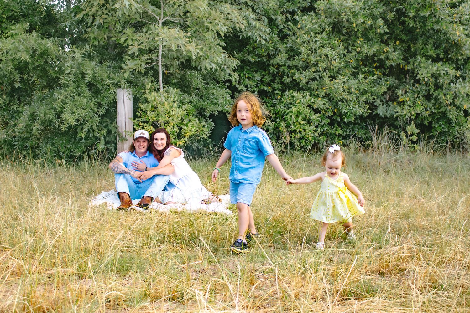 Family enjoying a sunny day outdoors, with two children playing in a grassy field while parents relax on a blanket.