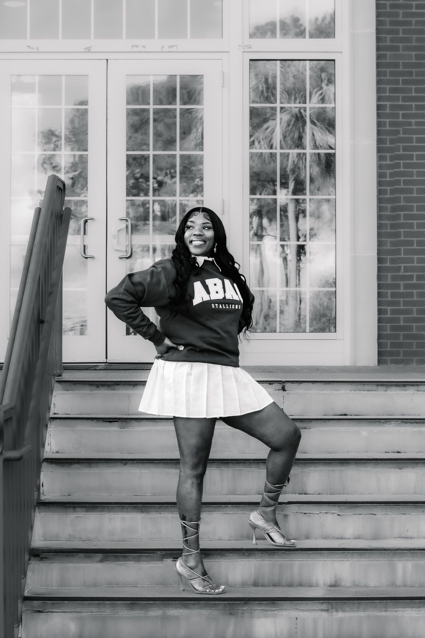 Young woman in ABAY sweatshirt and white pleated skirt poses confidently on brick building steps in black and white photo.
