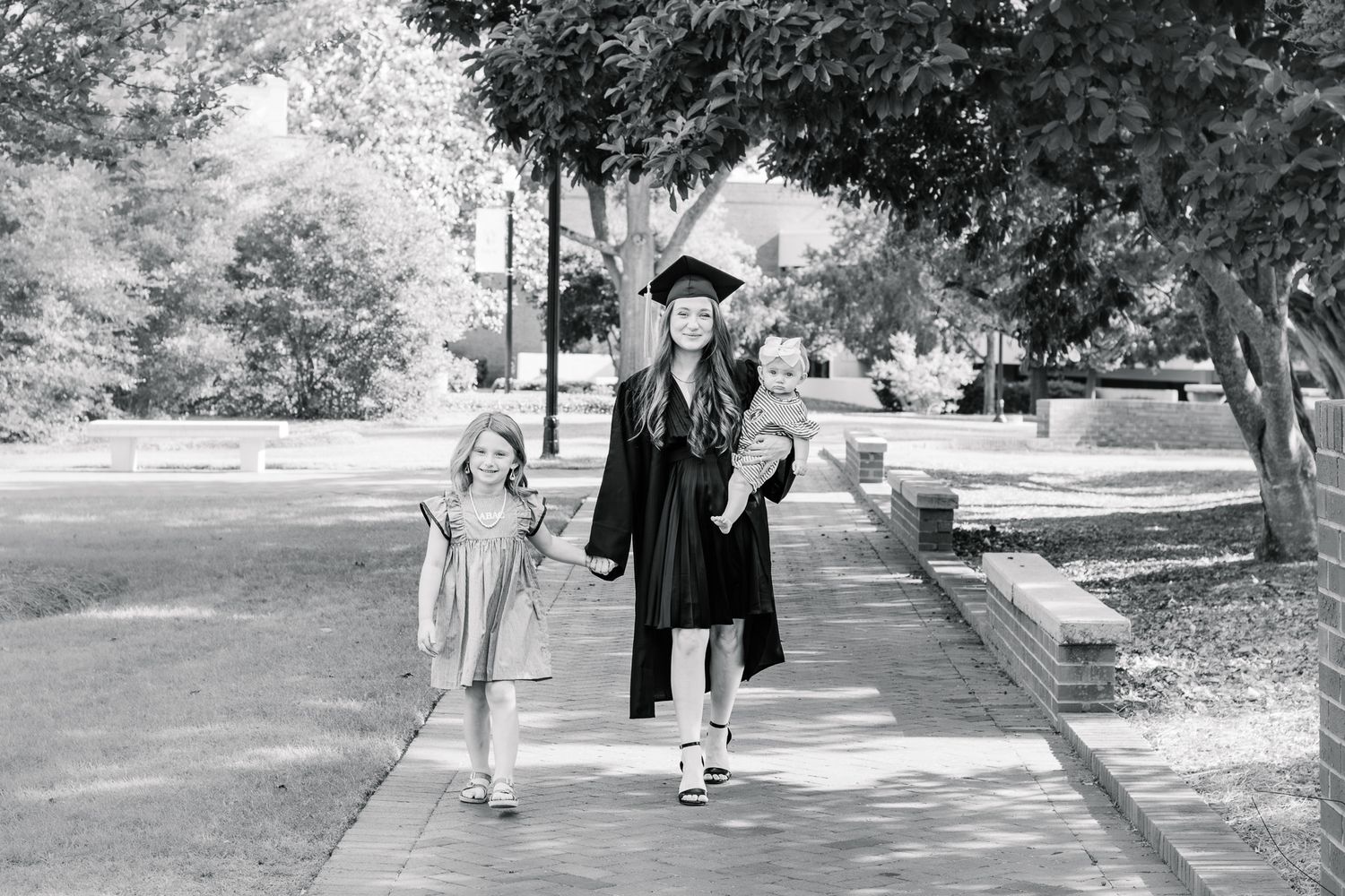 Graduate in cap and gown walks with two young children along tree-lined campus path in black and white.