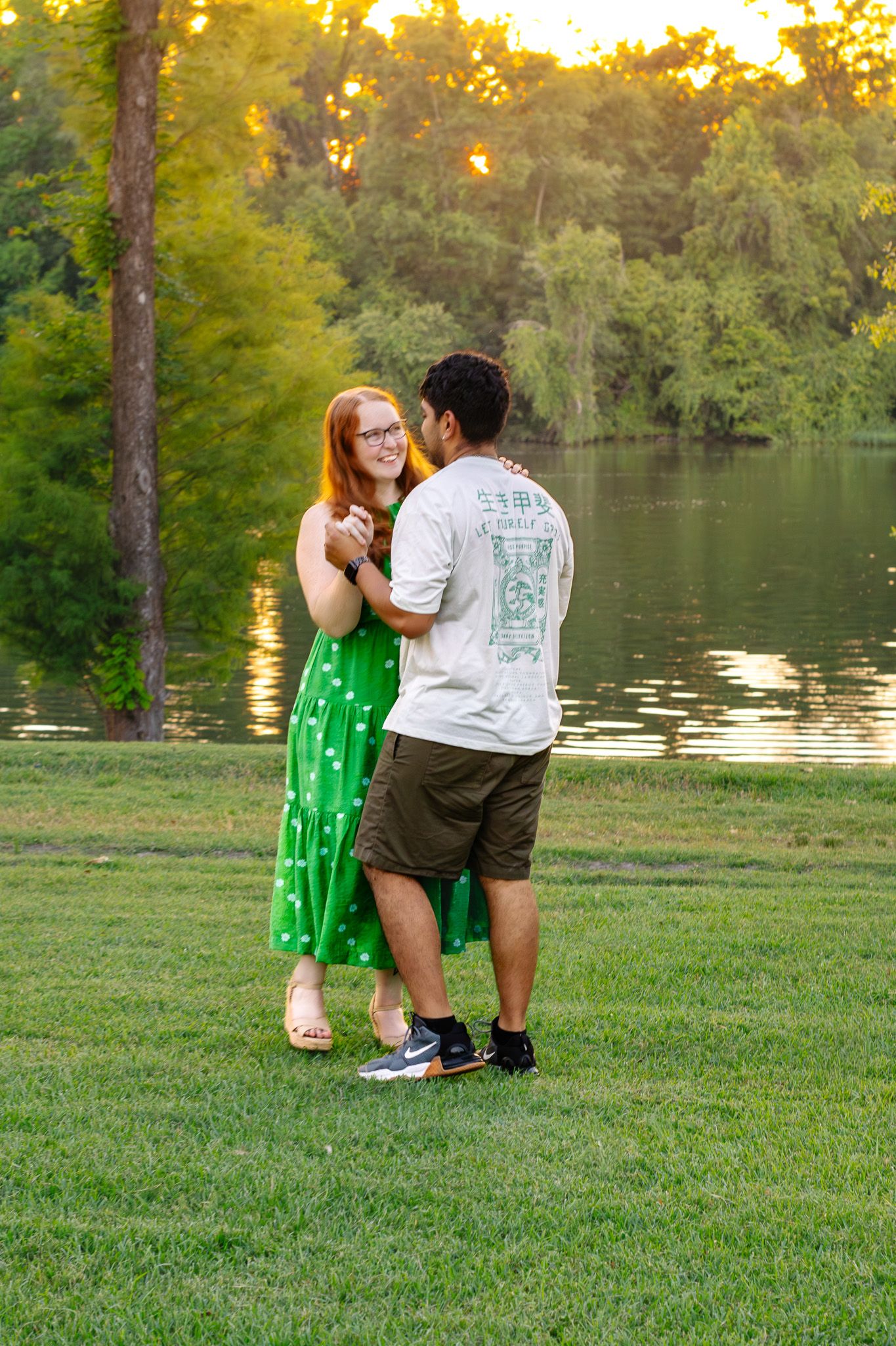 Couple dancing by a lake at sunset, woman in green polka dot dress, man in white graphic tee and khaki shorts.