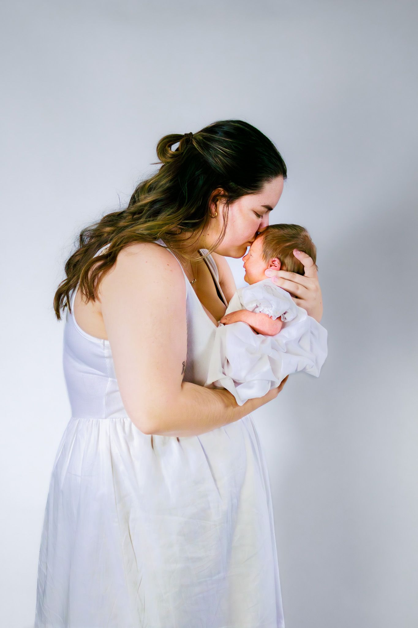 A loving mother in a white dress kissing her newborn baby held in her arms.
